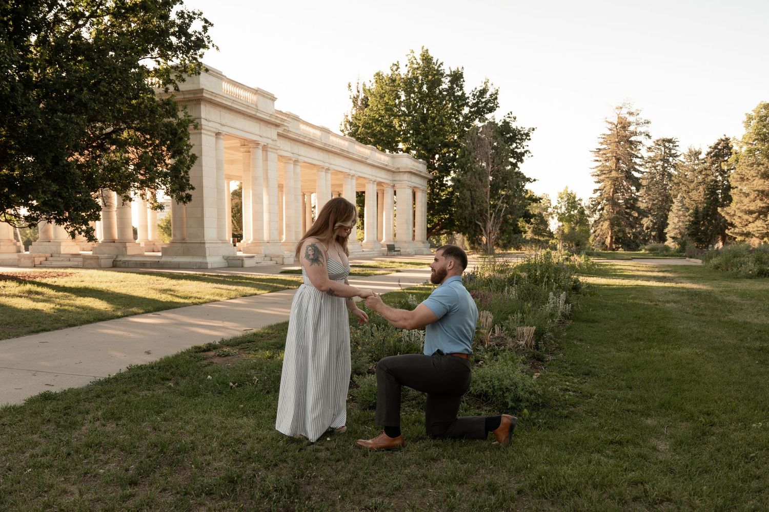 Man proposing on one knee to woman in white dress at golden hour near elegant stone pavilion in park.