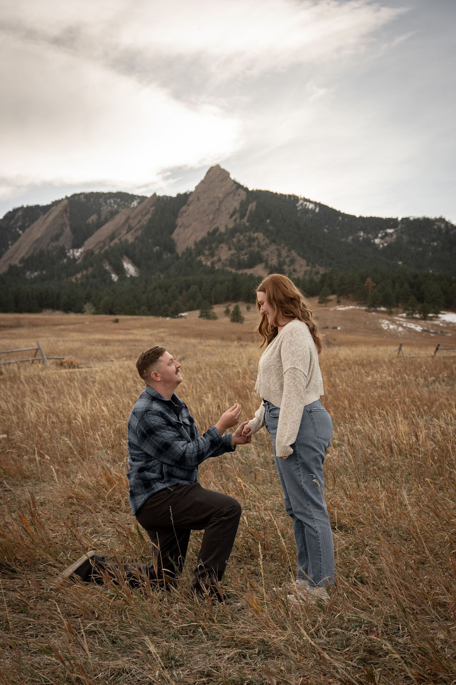 Man kneeling on one knee proposing to woman in golden field with Rocky Mountain Flatirons backdrop in Boulder, Colorado.