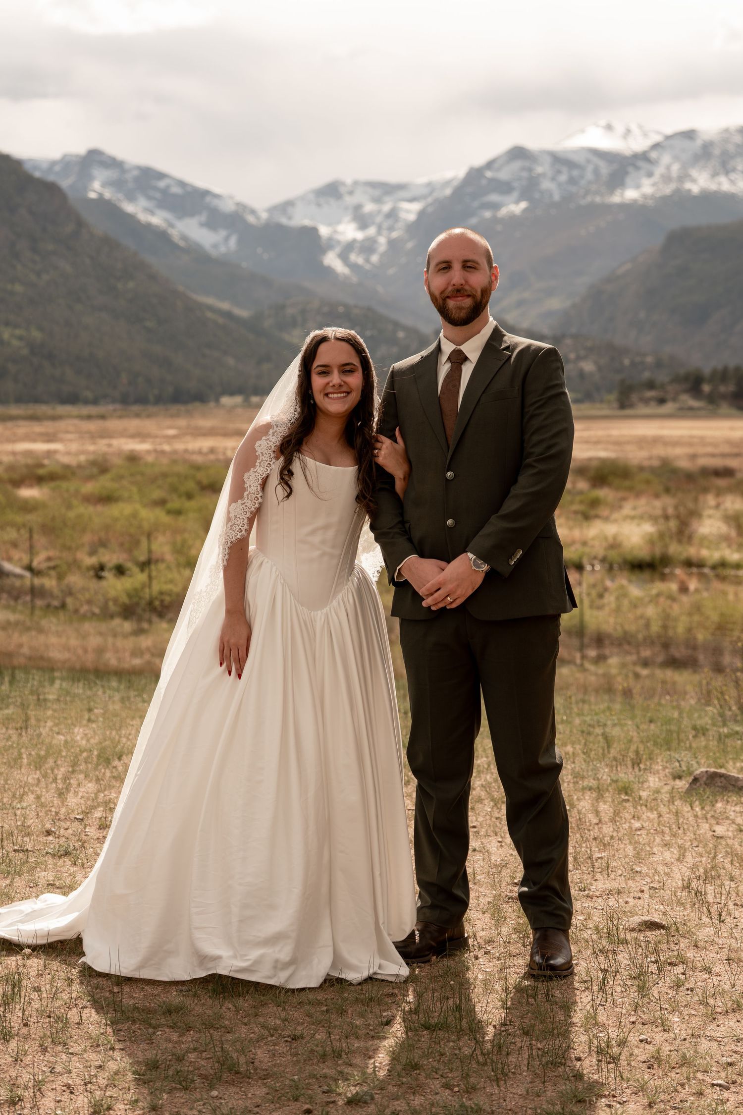 Smiling bride and groom pose together in front of stunning snow-capped mountain landscape.