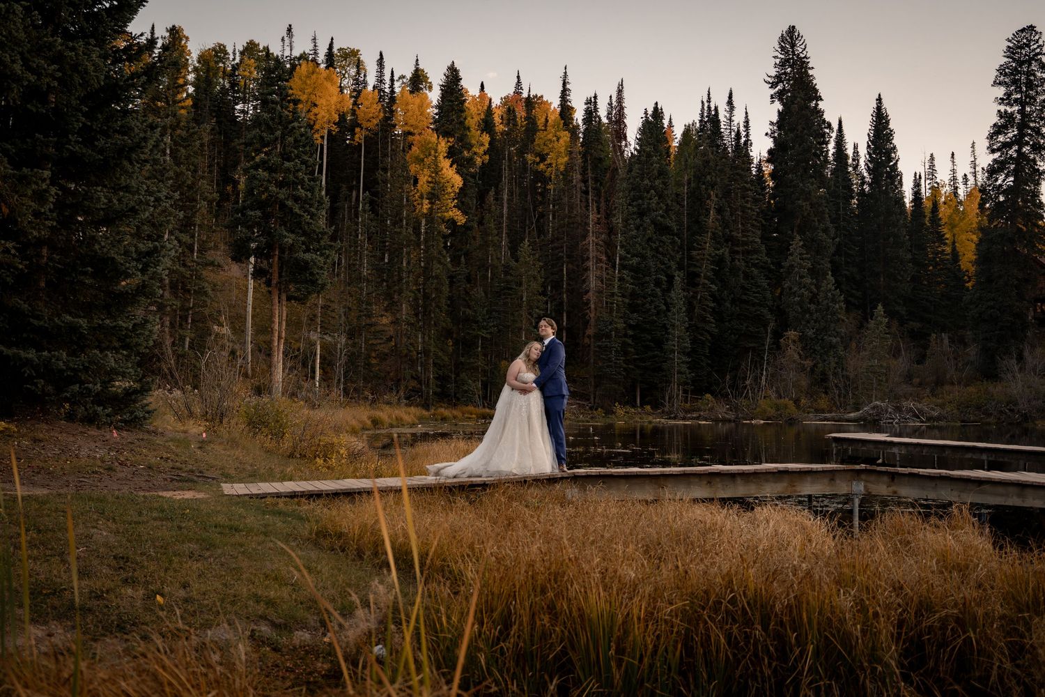 A couple stands on a wooden boardwalk against a backdrop of autumn-colored evergreen forest.