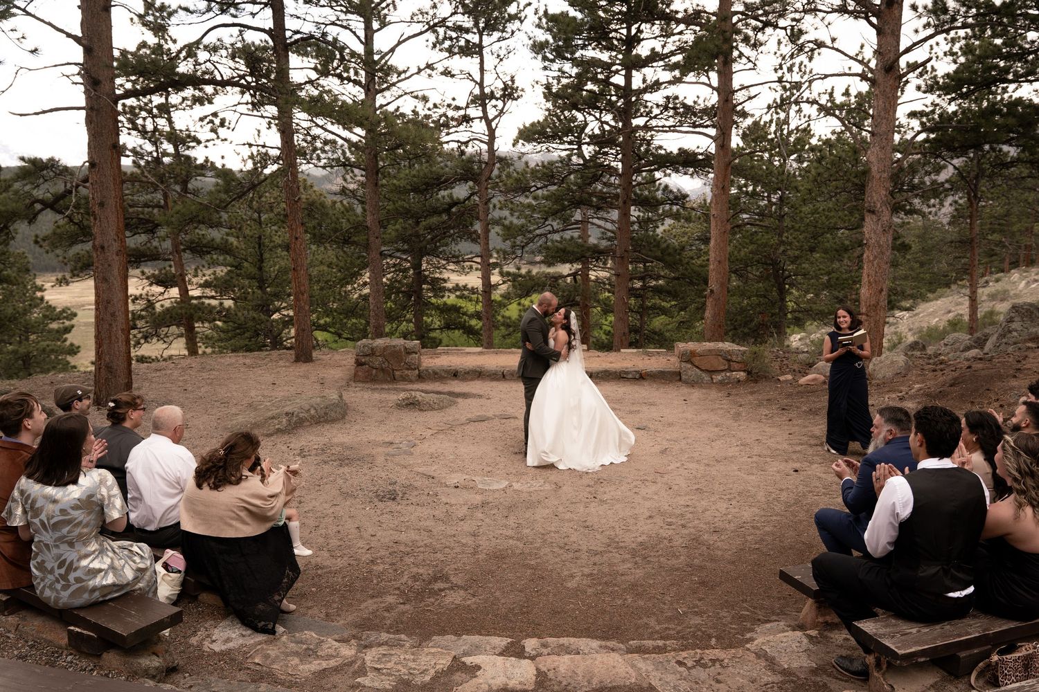 Outdoor wedding ceremony in a pine forest clearing with guests seated around the couple exchanging vows.