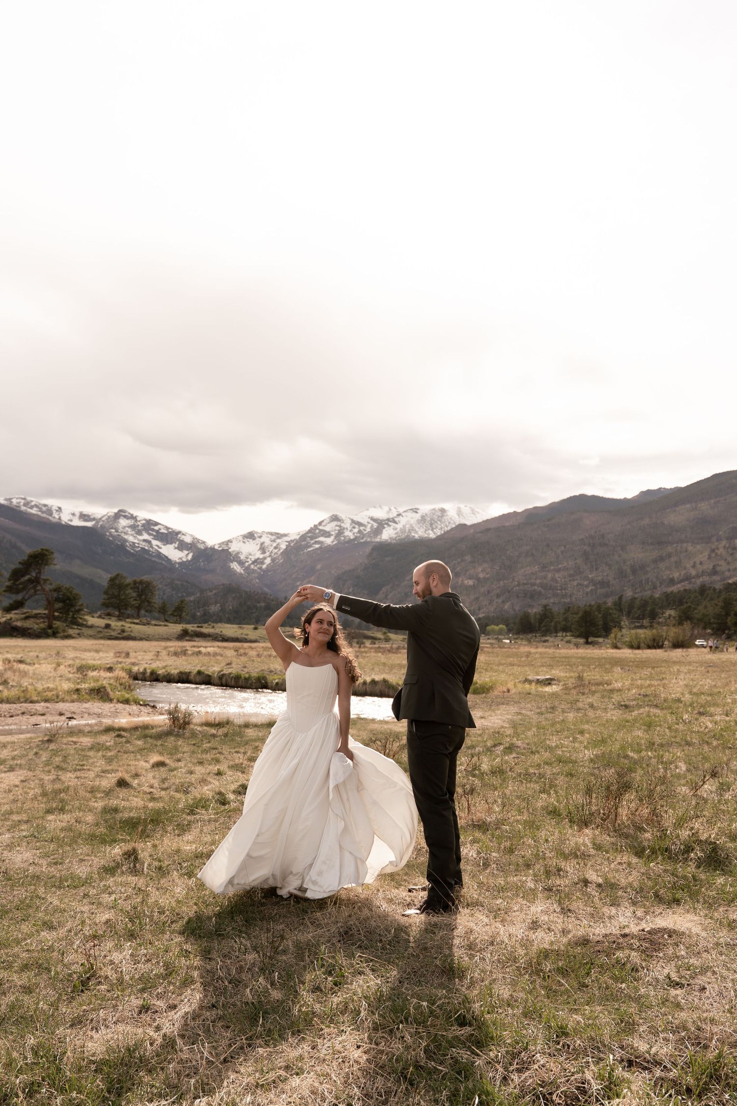 Bride and groom dancing in a mountain meadow, snow-capped peaks in the background, bride in flowing white gown.