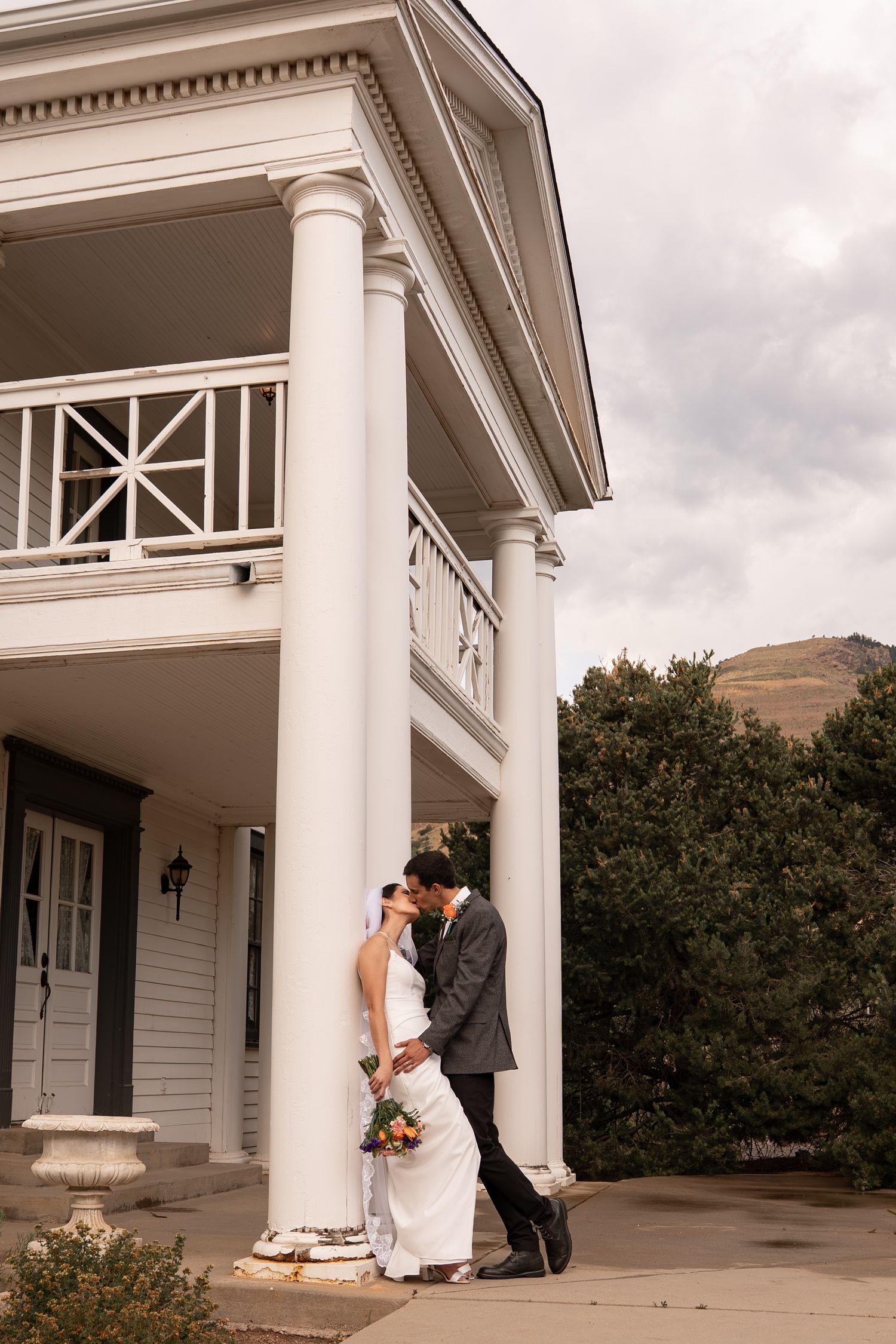 Bride and groom share a romantic kiss beside white columns of an elegant venue with mountain views.