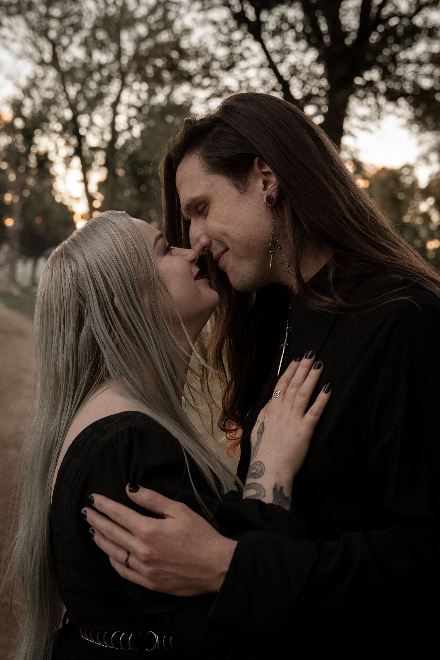A couple in black clothing shares a romantic kiss outdoors at sunset, surrounded by trees in a moody gothic setting.