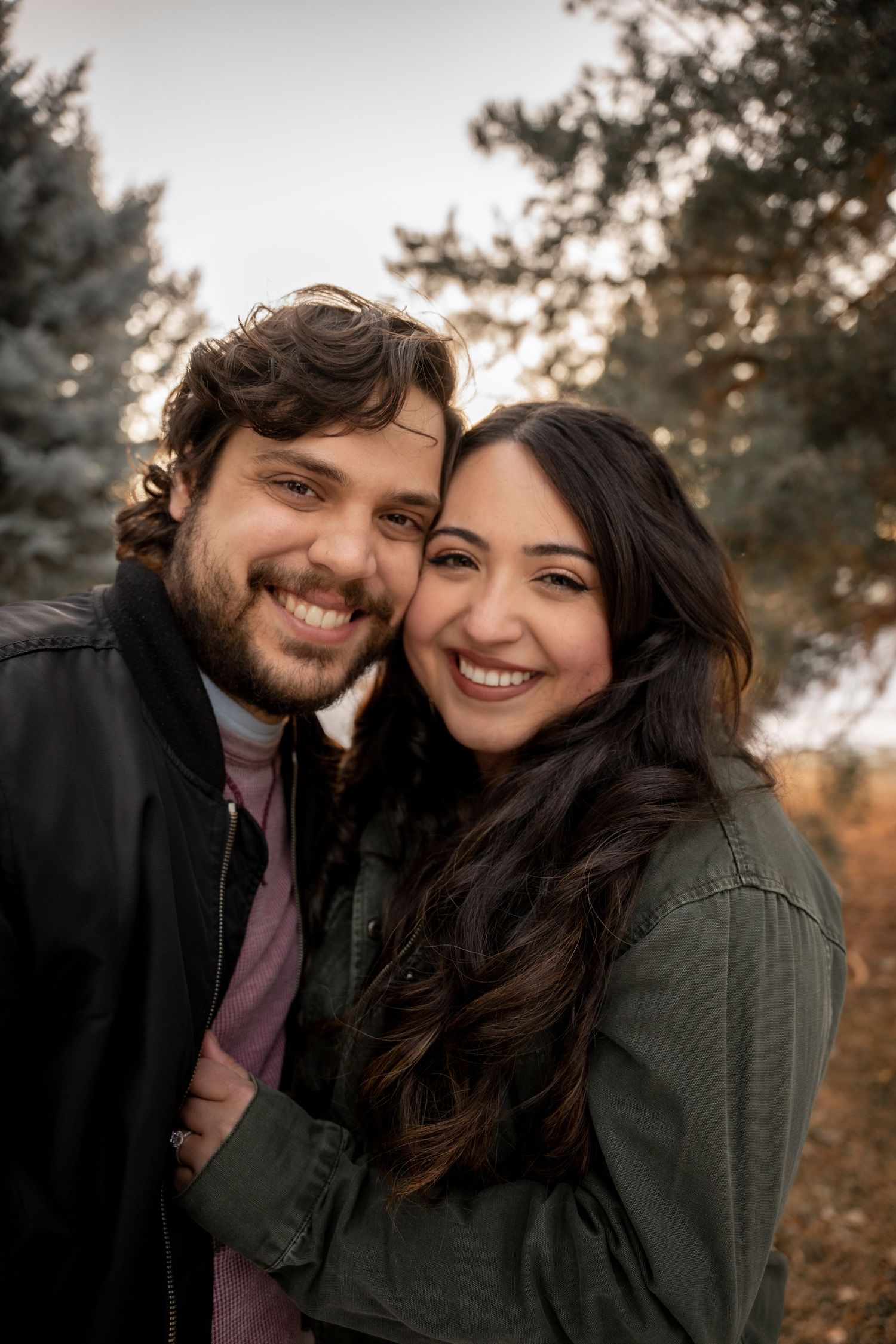 Smiling couple embracing outdoors among autumn trees, woman in green jacket leaning on man in black jacket.
