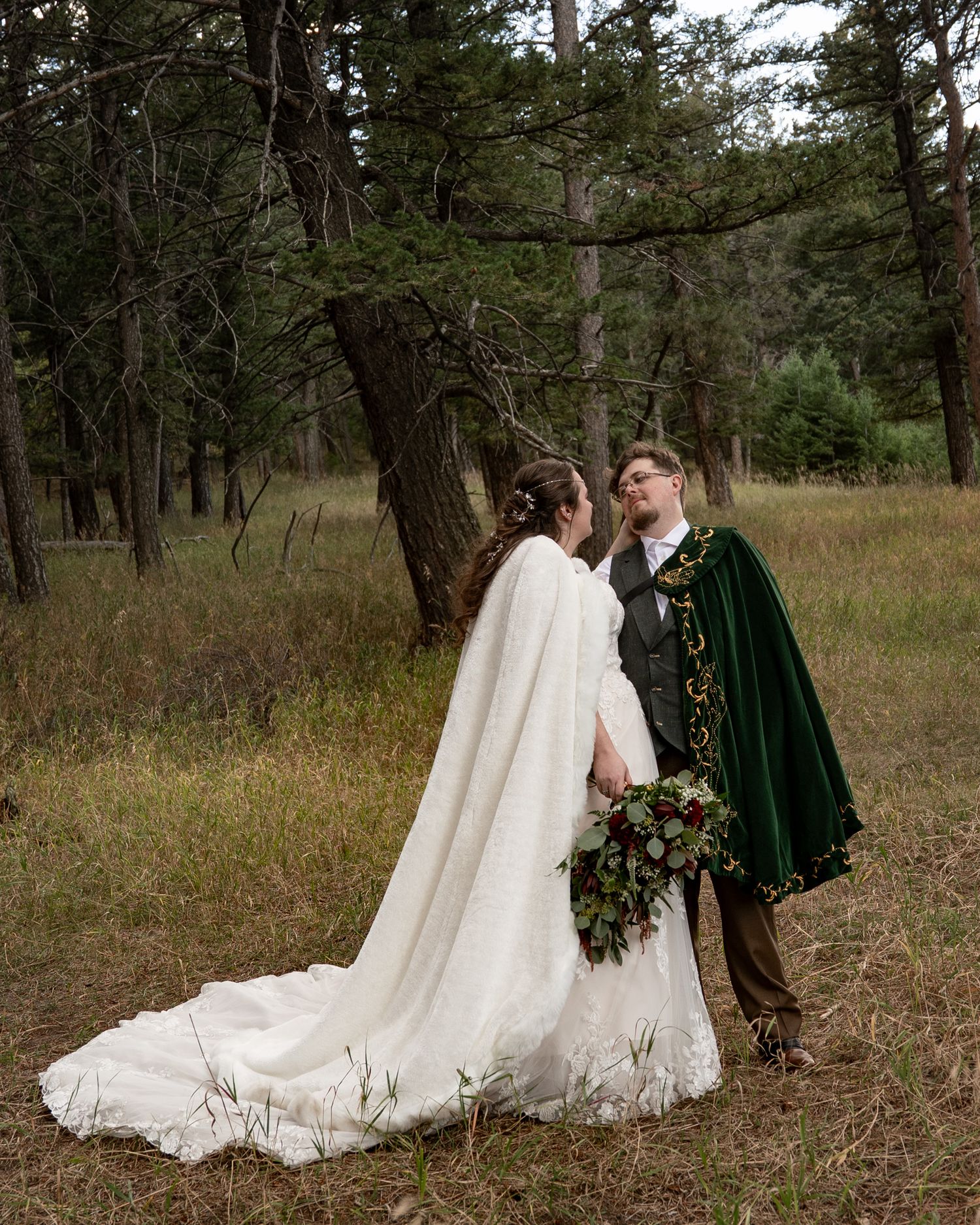Wedding couple in white dress and green cape photographed in a rustic forest setting.