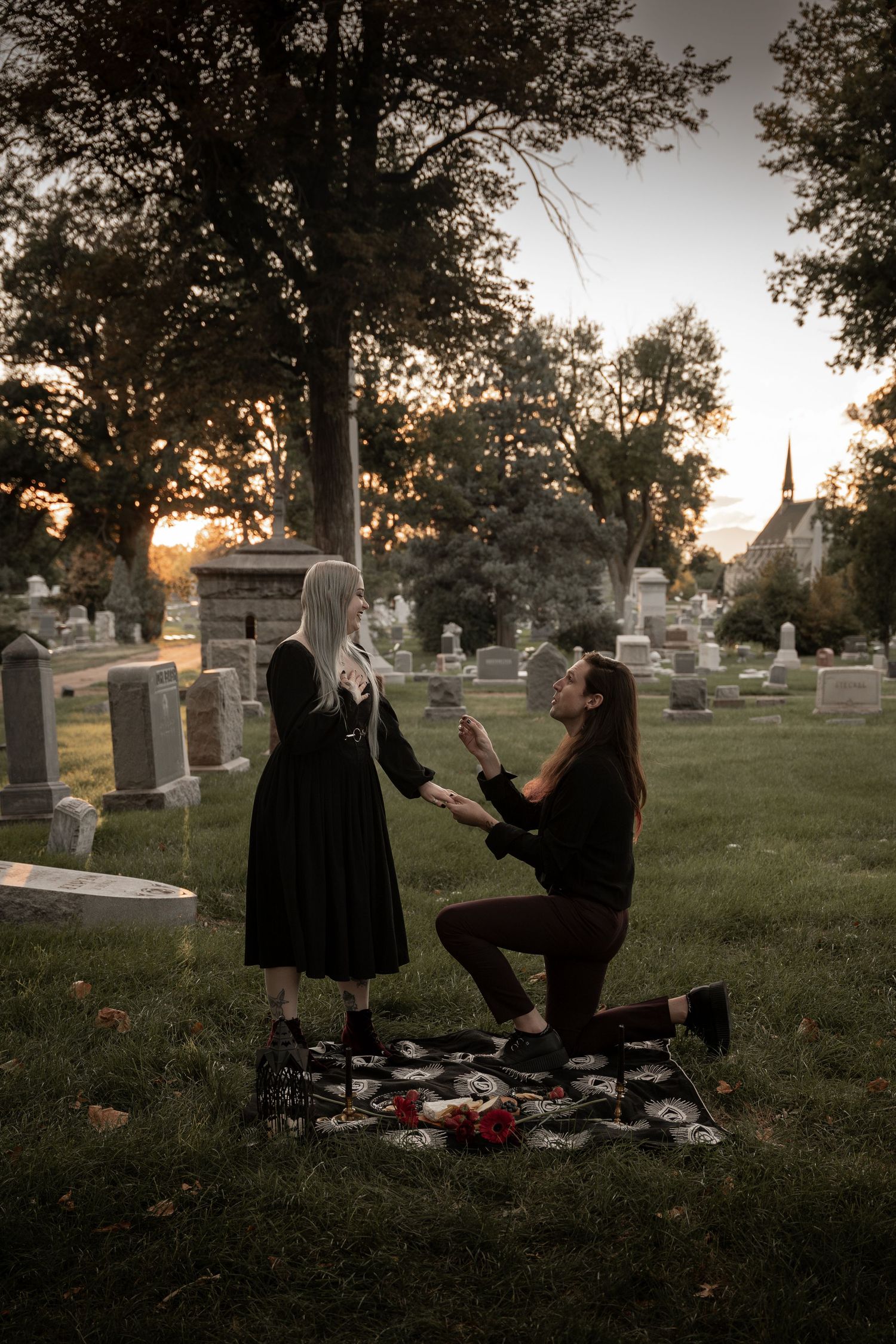 Couple sharing a romantic marriage proposal at sunset in a historic cemetery, with gravestones and golden light in background.