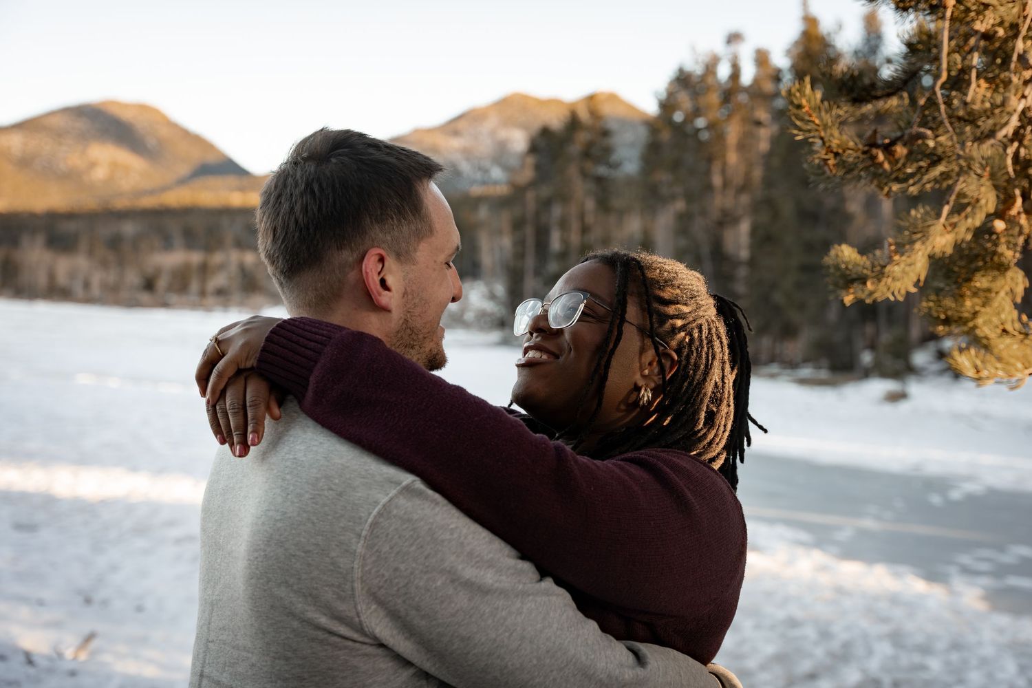 Interracial couple embracing outdoors in winter, snowy mountain landscape with pine trees in background.