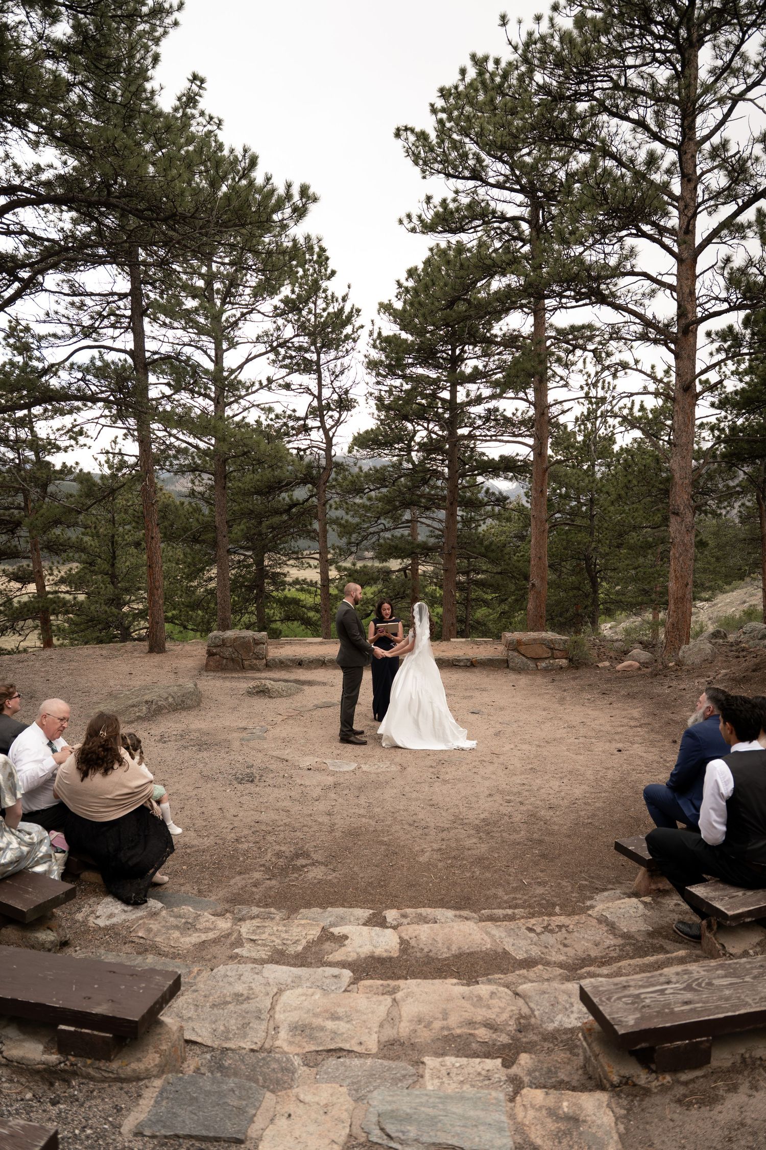 Outdoor wedding ceremony in pine forest amphitheater in Moraine Park with couple at altar and guests seated on stone benches.