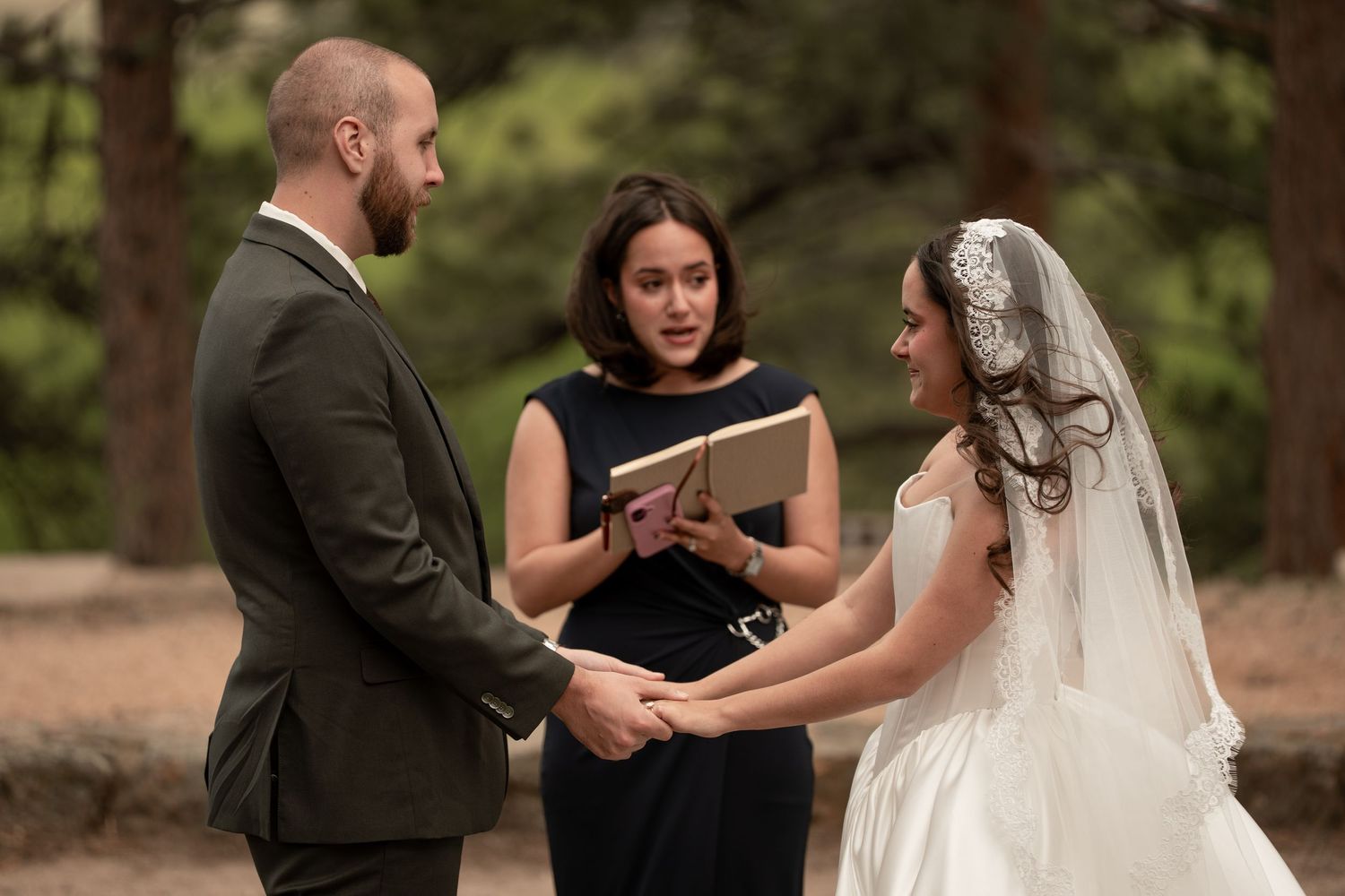 Outdoor wedding ceremony with bride in white gown and veil, groom in suit, holding hands before officiant in forest.