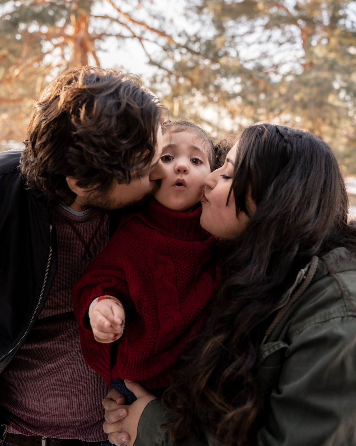 Two parents kissing their toddler on the cheeks in a warm outdoor family portrait among trees.