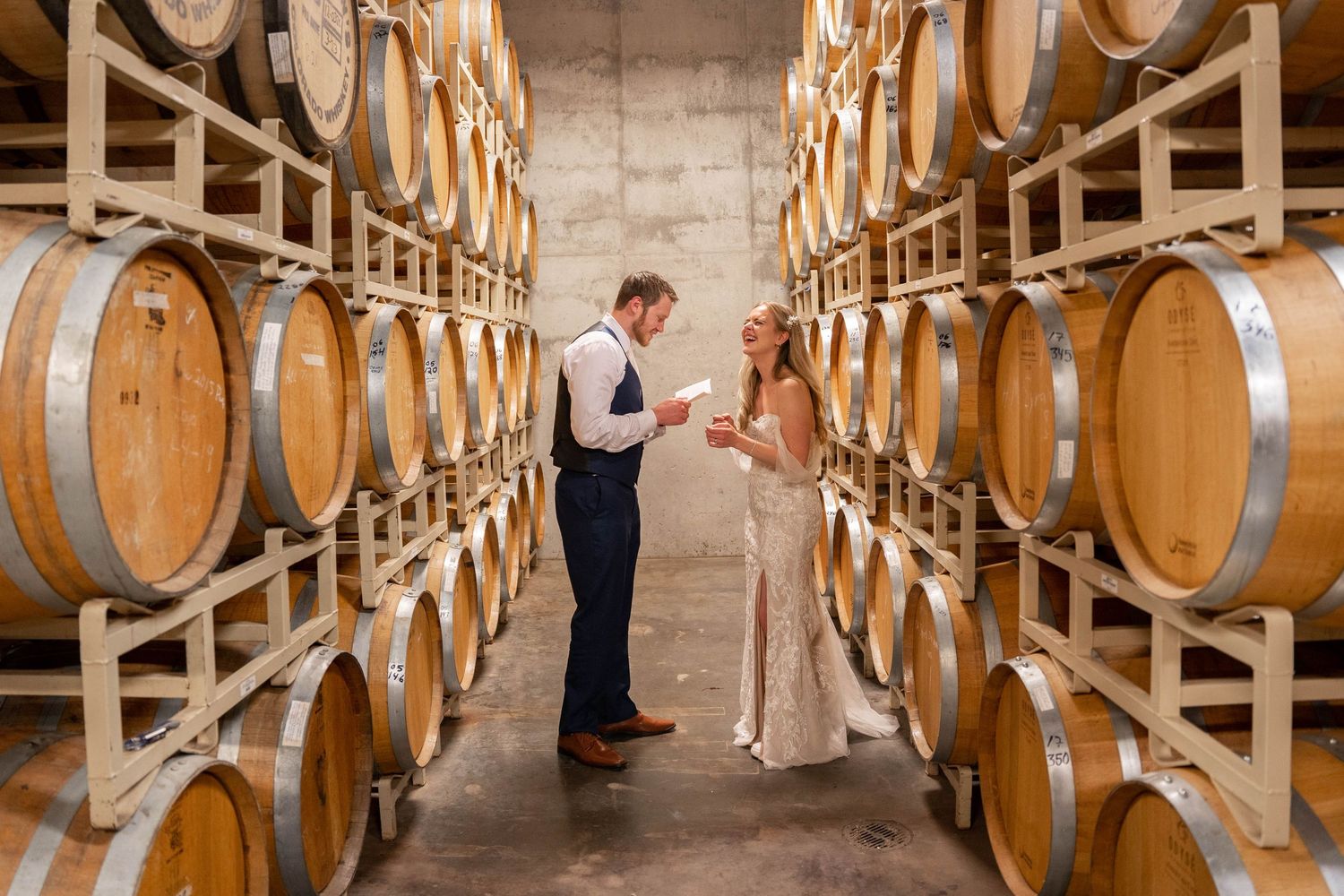 Rows of wooden wine barrels line both sides of a long cellar corridor with stone walls and concrete floor.