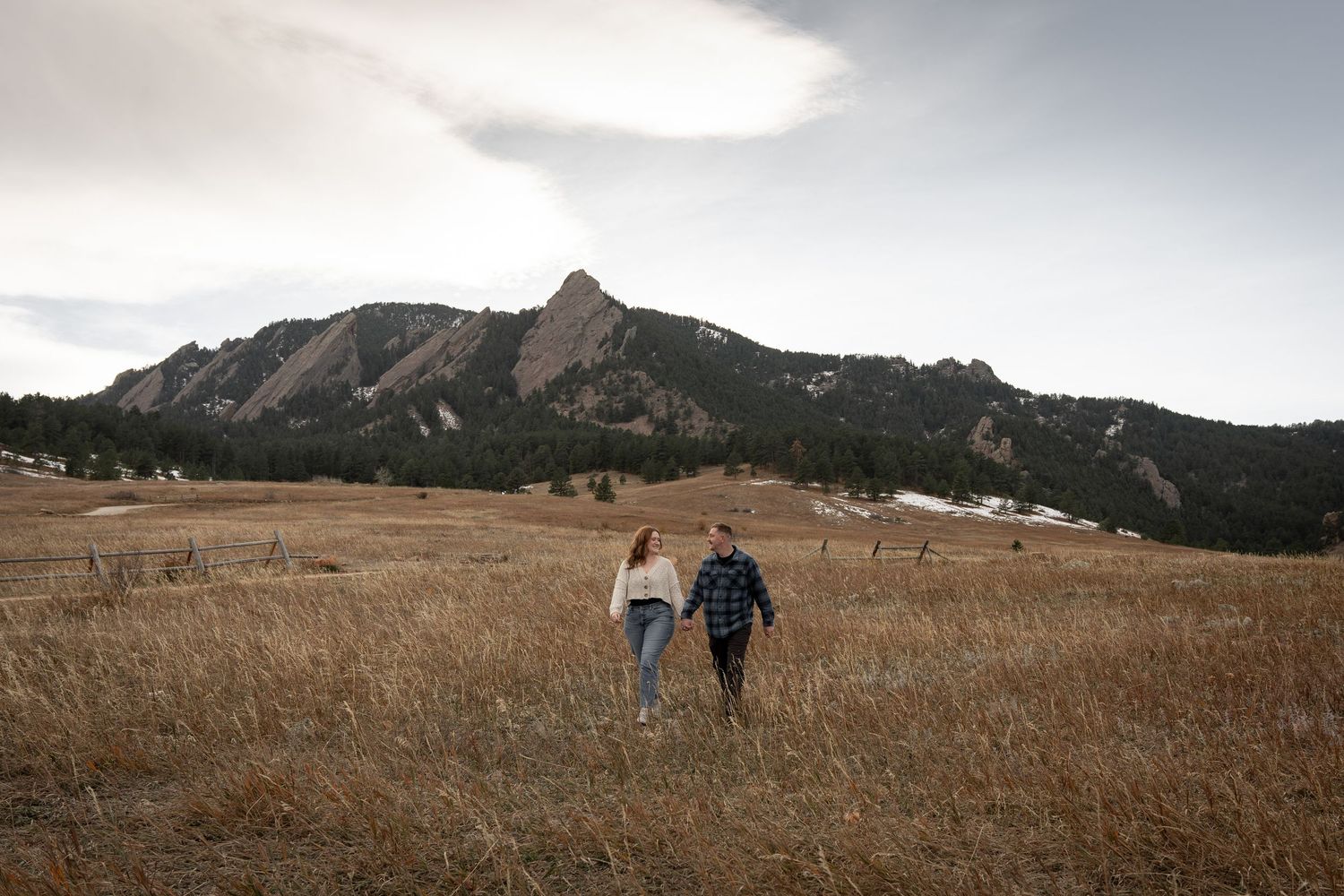 Couple holding hands walking through golden meadow with dramatic Flatirons rock formations in Boulder, Colorado backdrop.