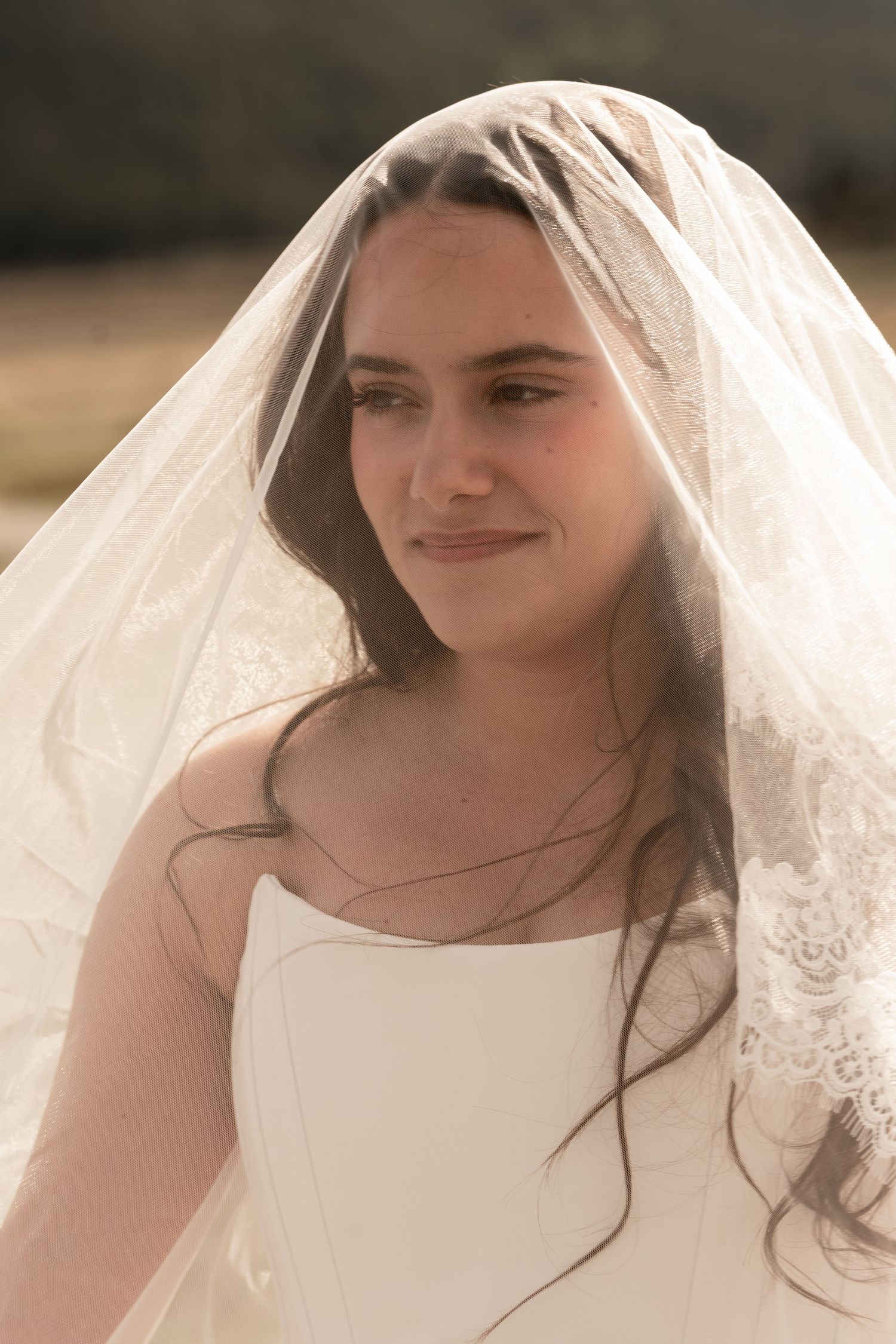 Beautiful bride smiles softly beneath a delicate lace wedding veil in a sun-drenched outdoor setting.