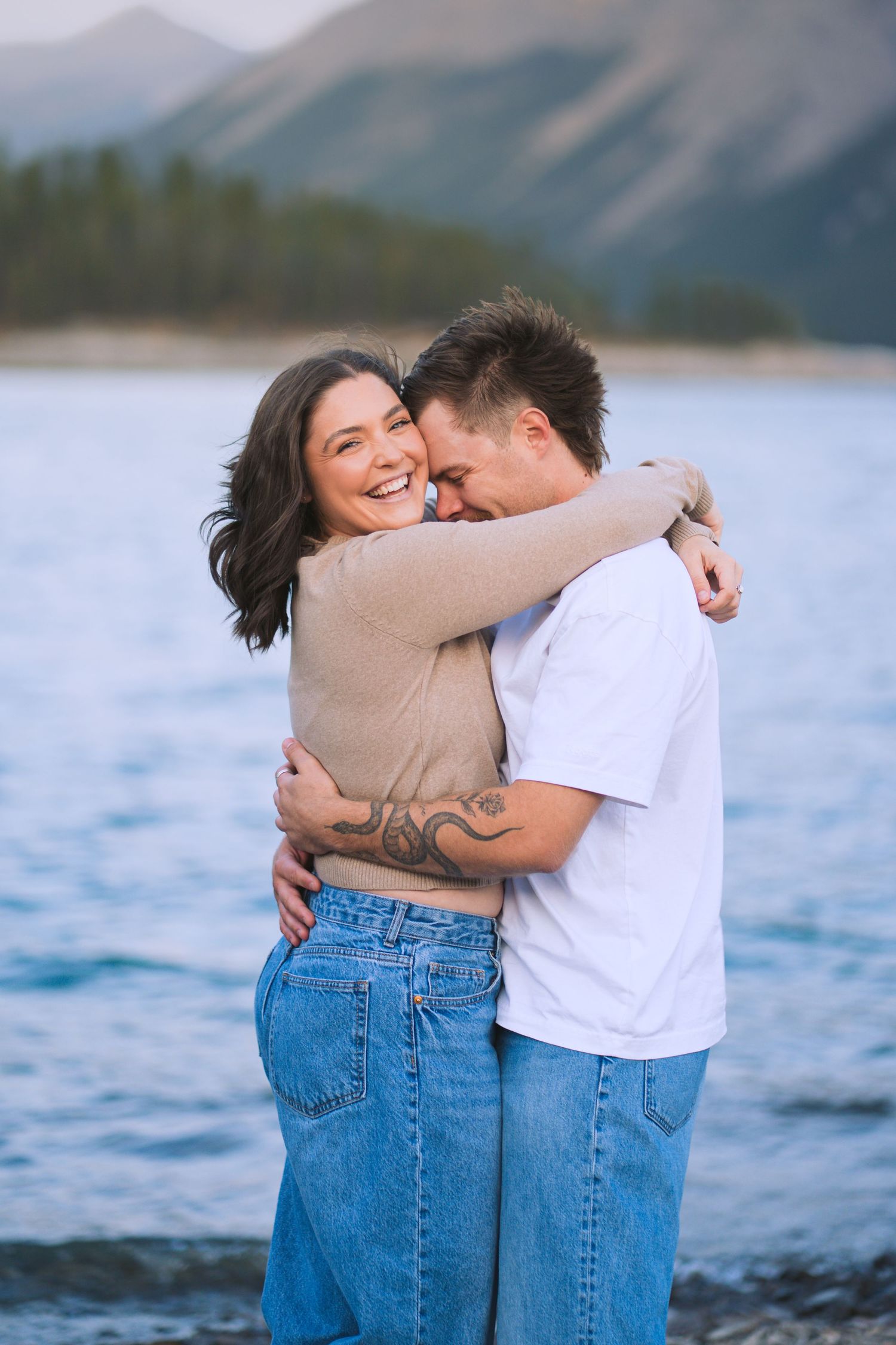 Couple holding each other closely at Lake Minnewanka in Banff, sharing an intimate moment with mountains and water behind them.