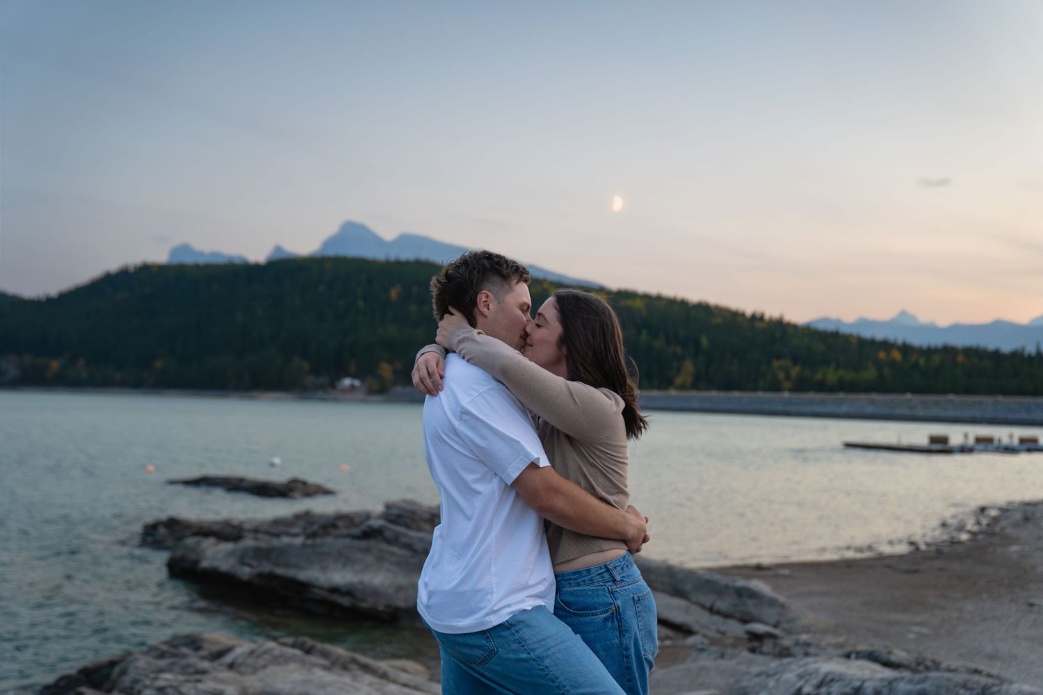 Couple share a kiss at Lake Minnewanka in Banff, with mountains and calm water at dusk, capturing a romantic and natural engagement moment.
