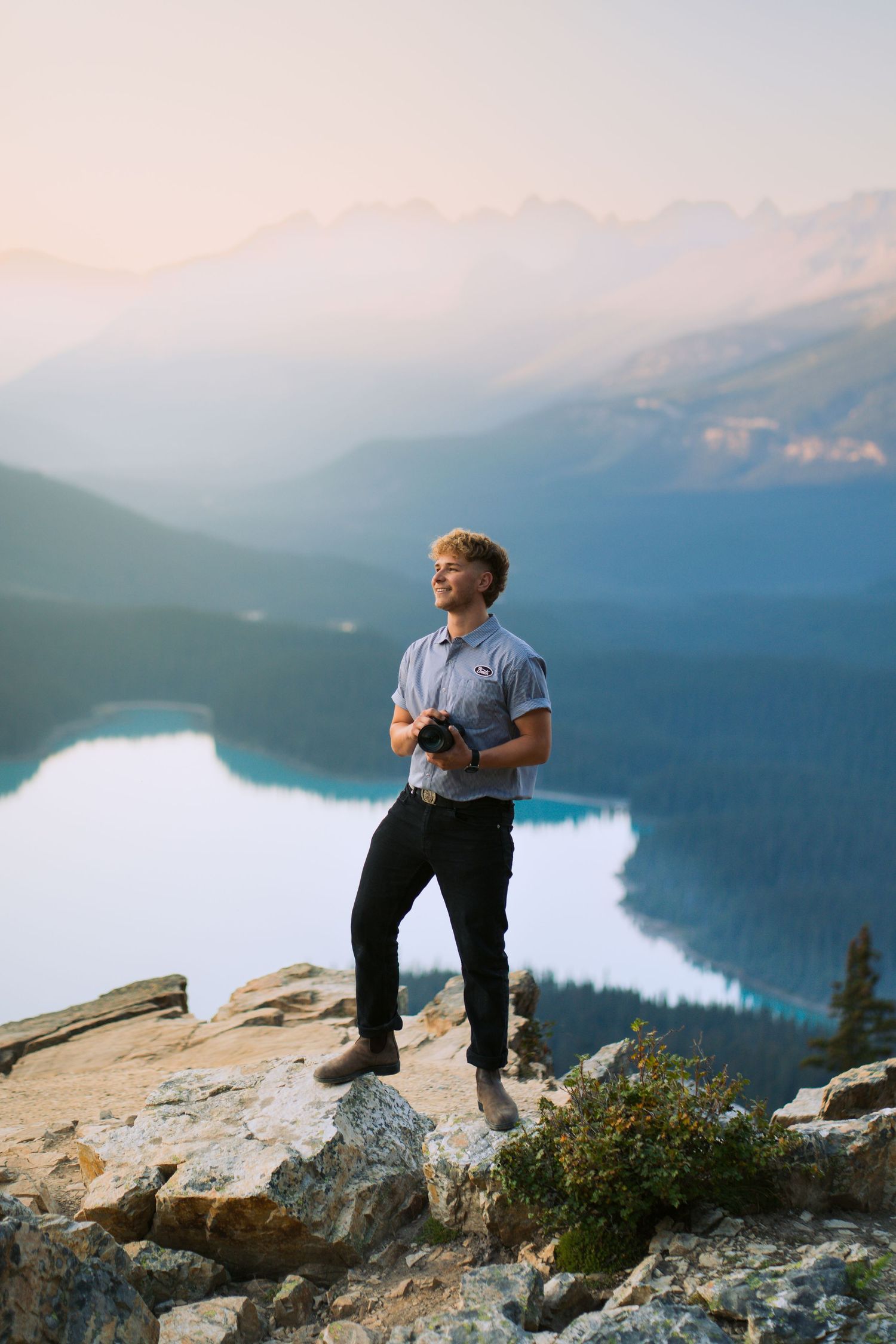A photographer stands on a rocky overlook above a bright blue alpine lake at sunset in Banff National Park, holding a camera and looking off