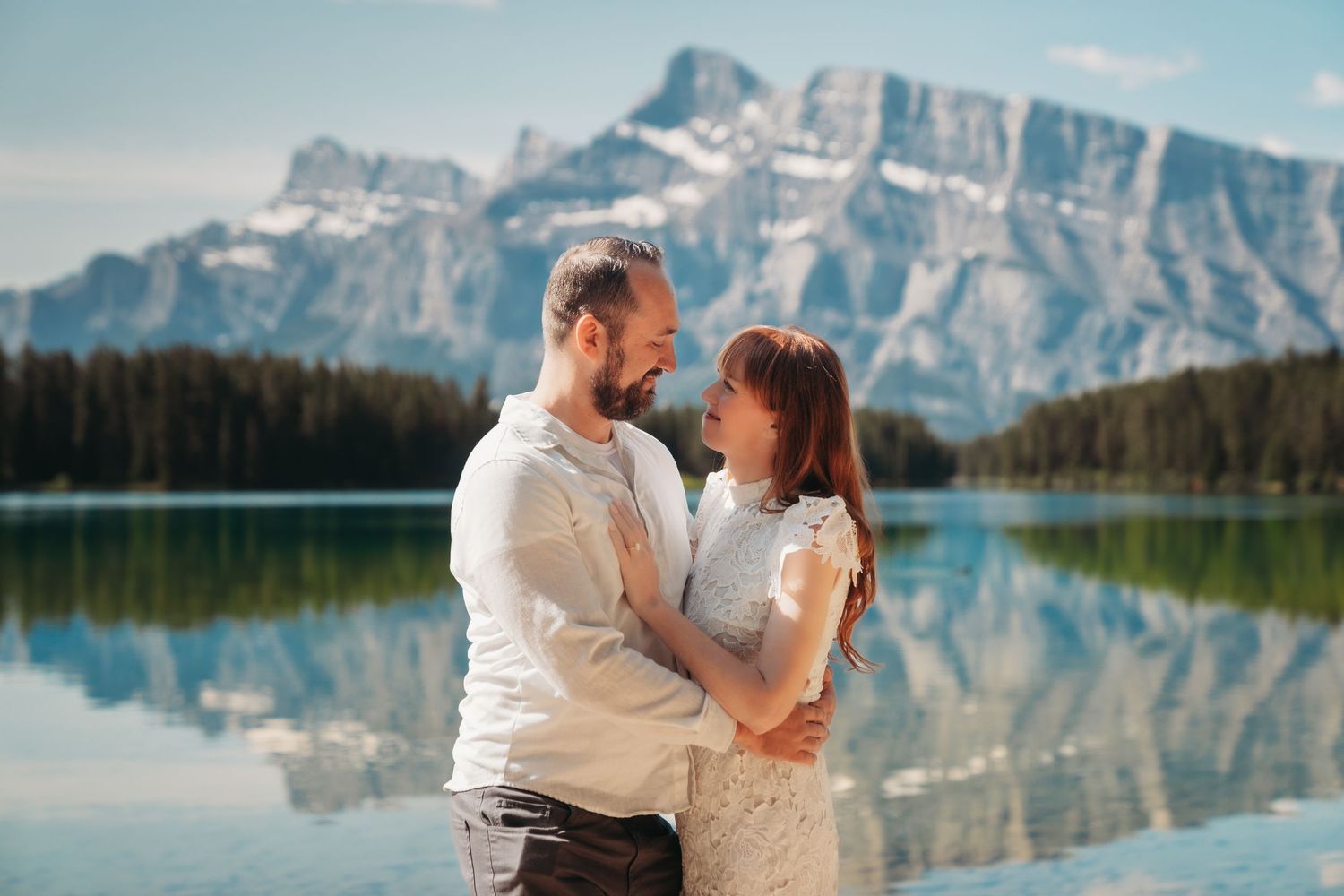 Bride and groom stand smiling by a calm mountain lake in Banff, surrounded by alpine peaks, evergreen trees, and clear blue summer skies.