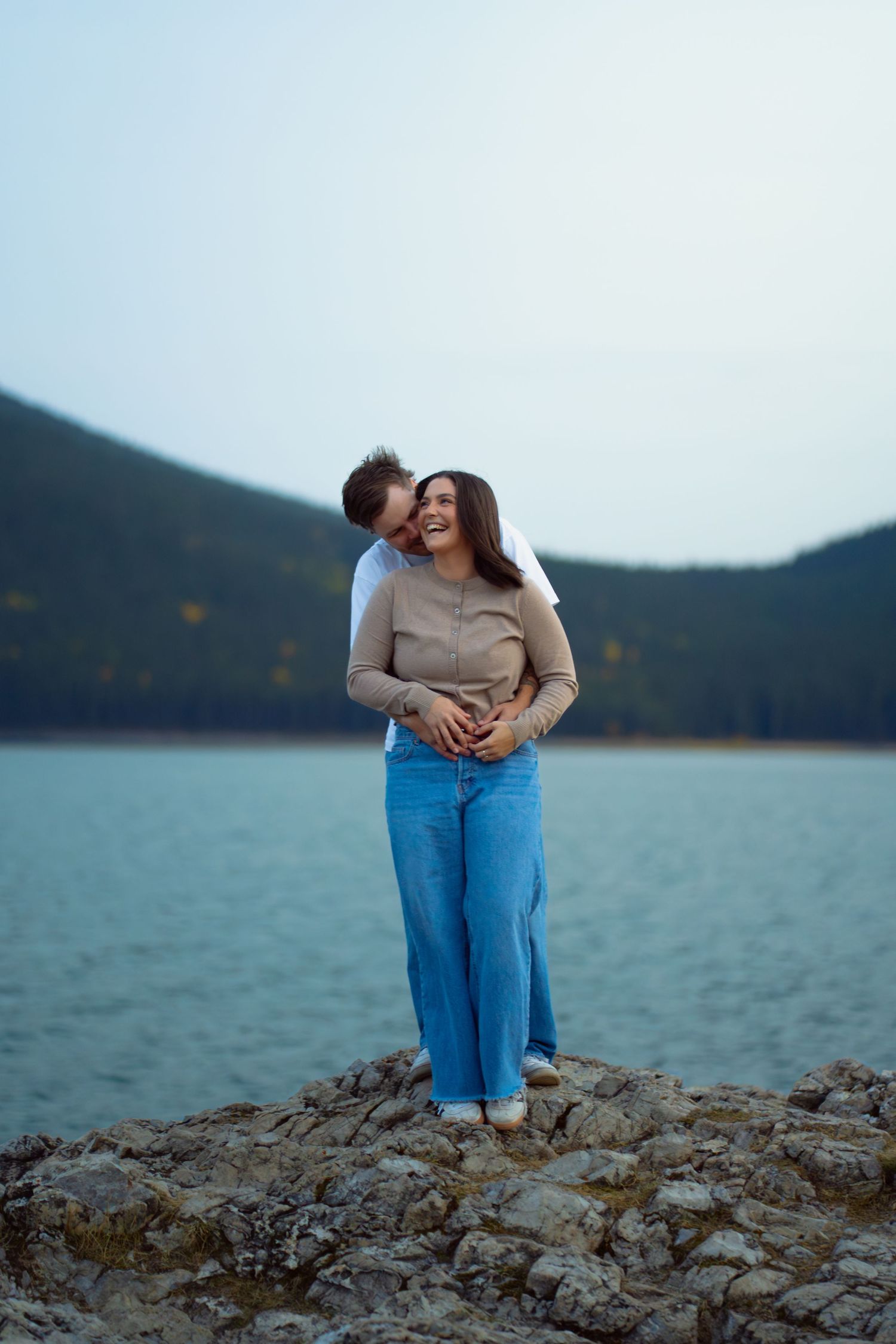 Couple hugging and laughing on the rocky shore at Lake Minnewanka in Banff, with water and forested mountains in the background.