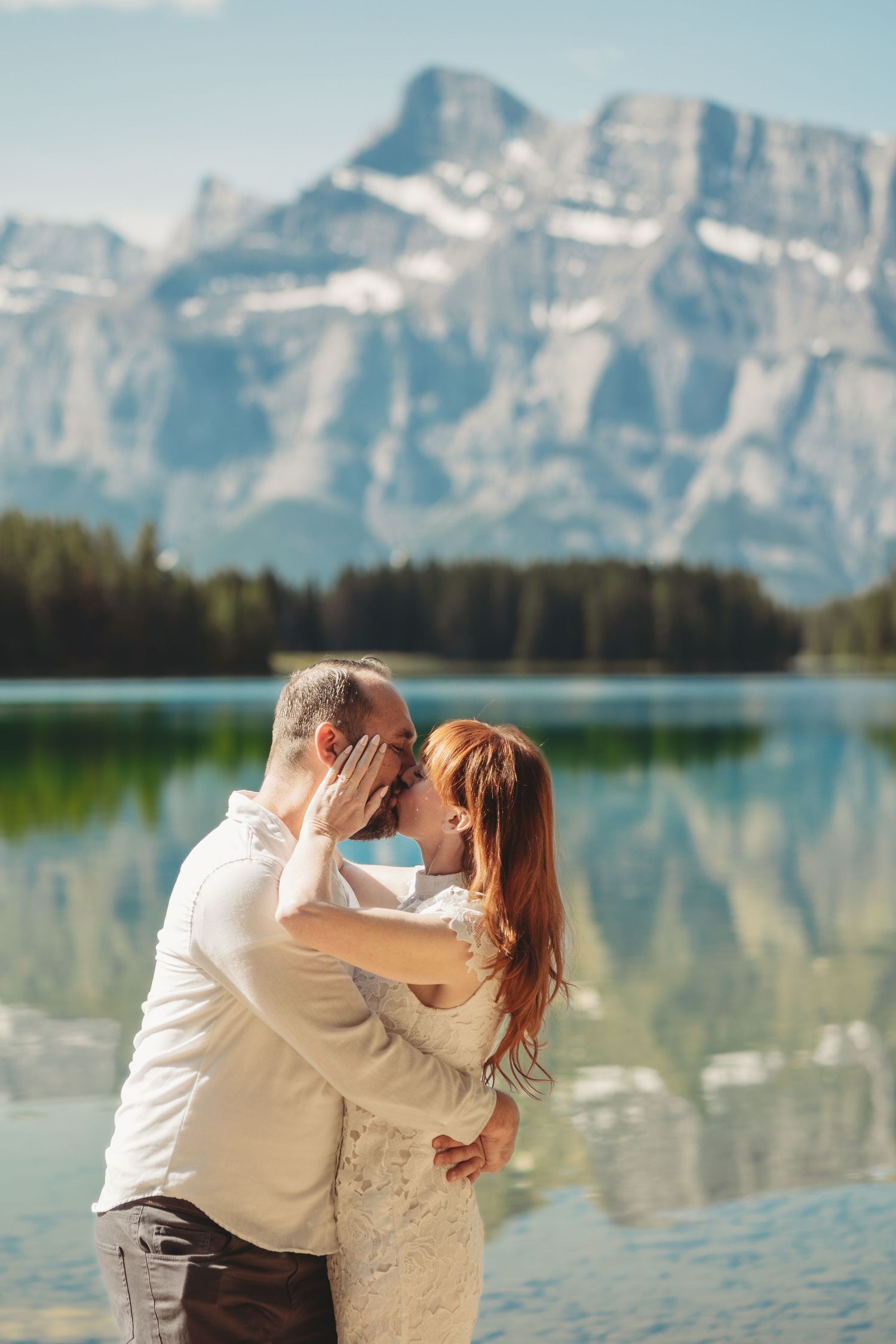 Bride and groom stand together sharing a kiss at 2 jack lake in Banff with crystal reflections and dramatic Rocky Mountain peaks.