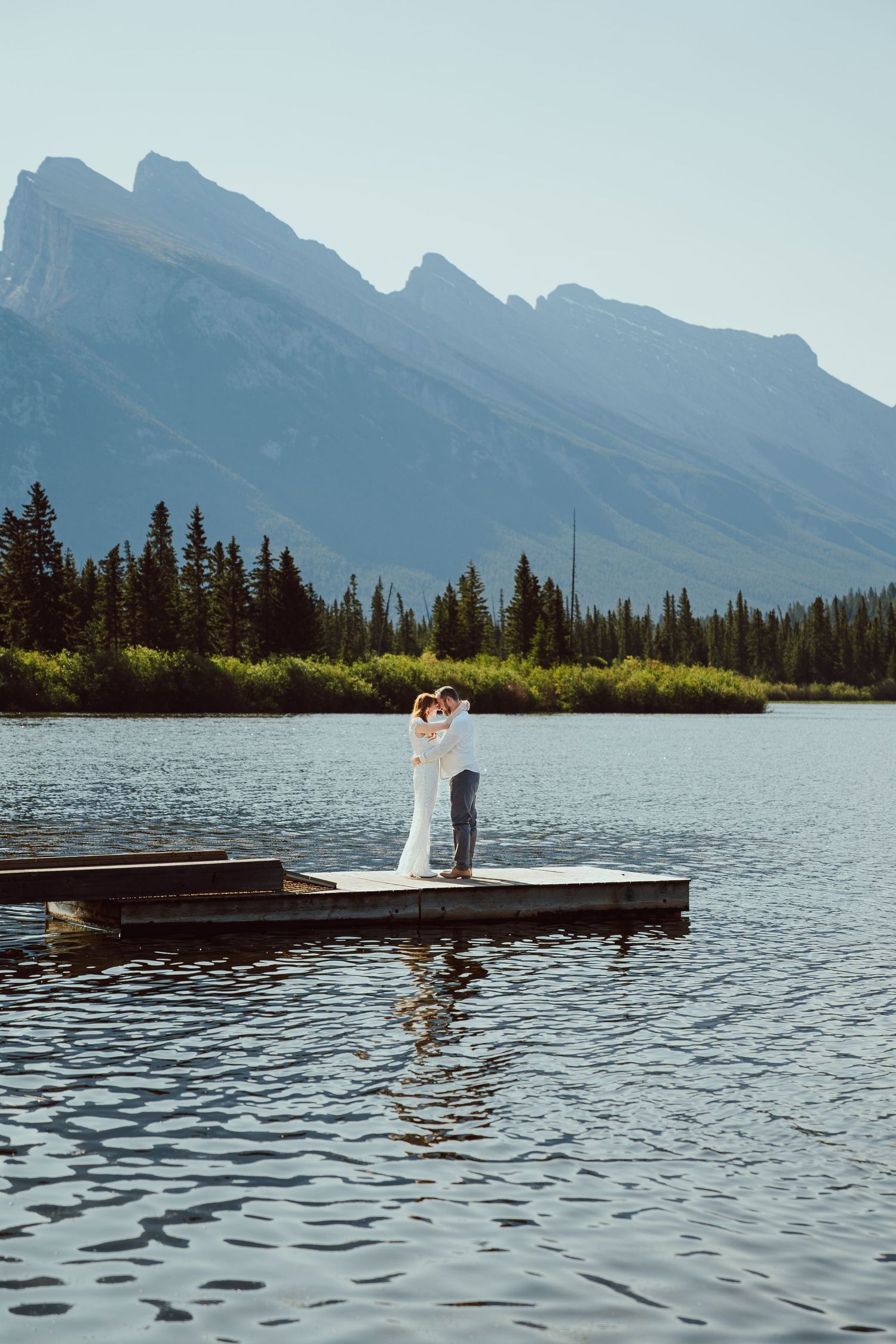 Bride and groom embrace on a dock at a mountain lake in Banff, surrounded by calm water, evergreen trees, and dramatic Rocky Mountain peaks.