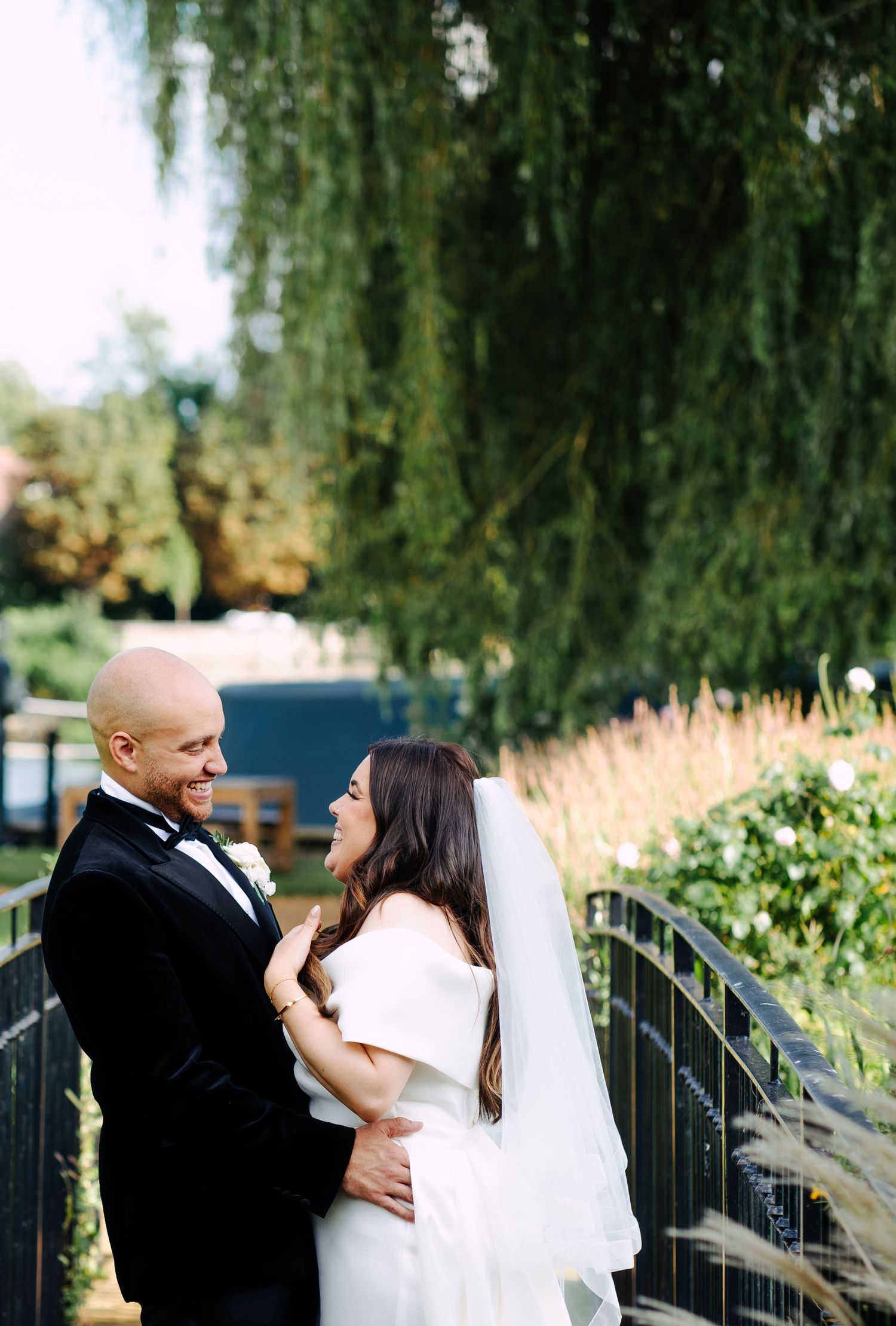 A joyful wedding portrait of a newly married couple standing together on a bridge, smiling directly at the camera.