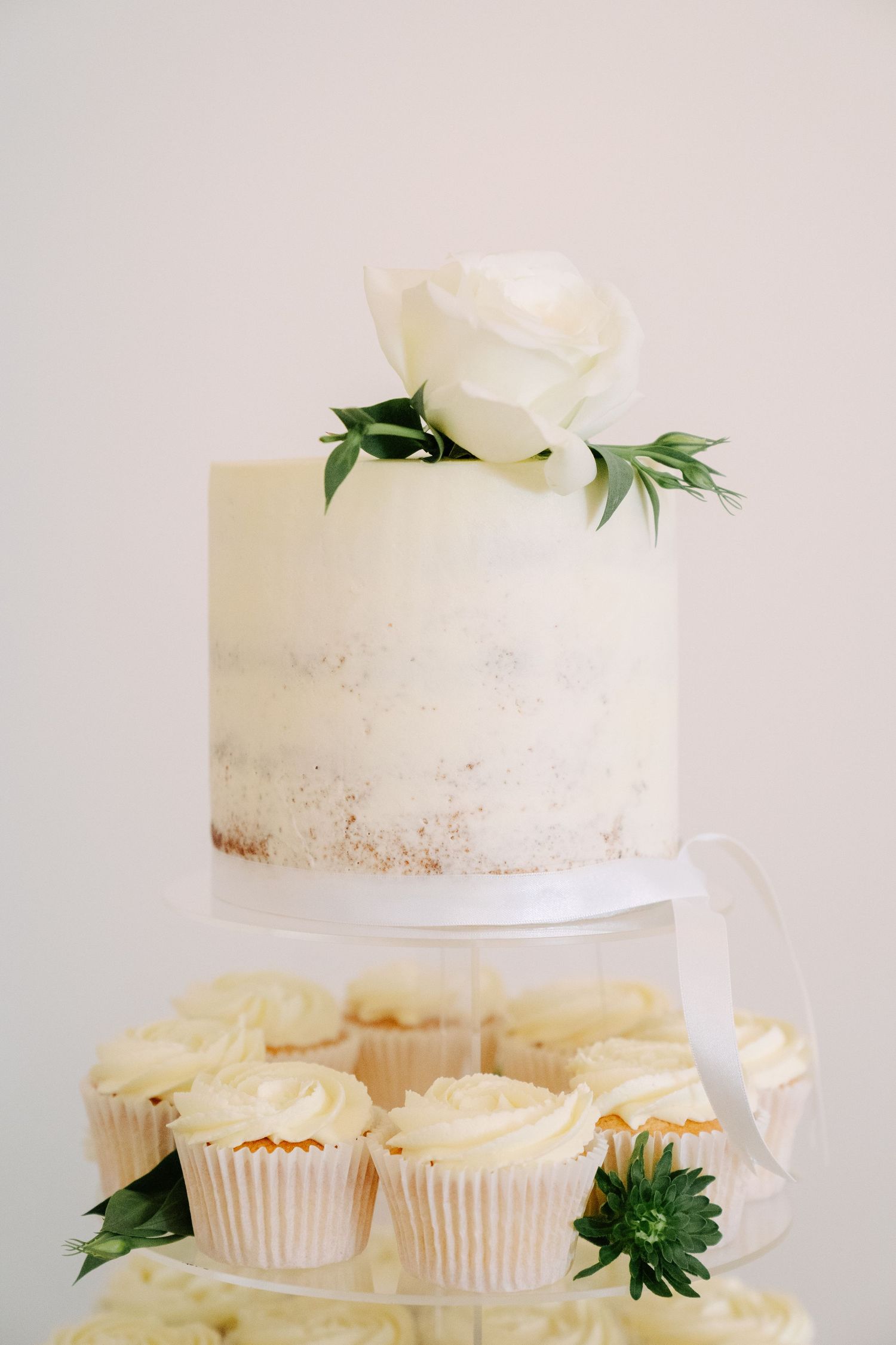 White semi-naked wedding cake topped with fresh roses, surrounded by matching frosted cupcakes on a clear display stand.
