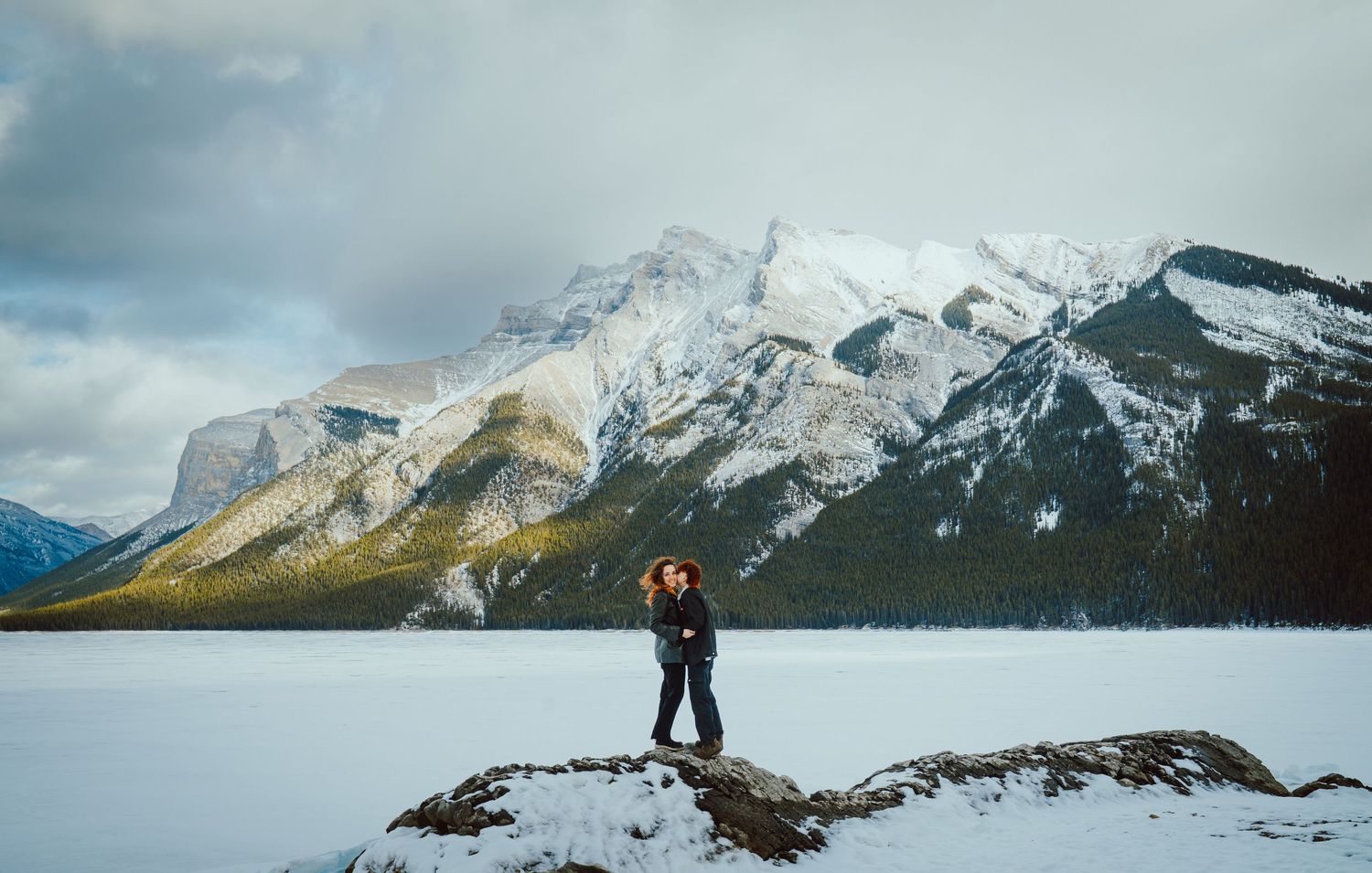 Couple sharing a kiss on a snowy lakeshore with dramatic Rocky Mountain peaks behind them, capturing a romantic winter moment in Banff.