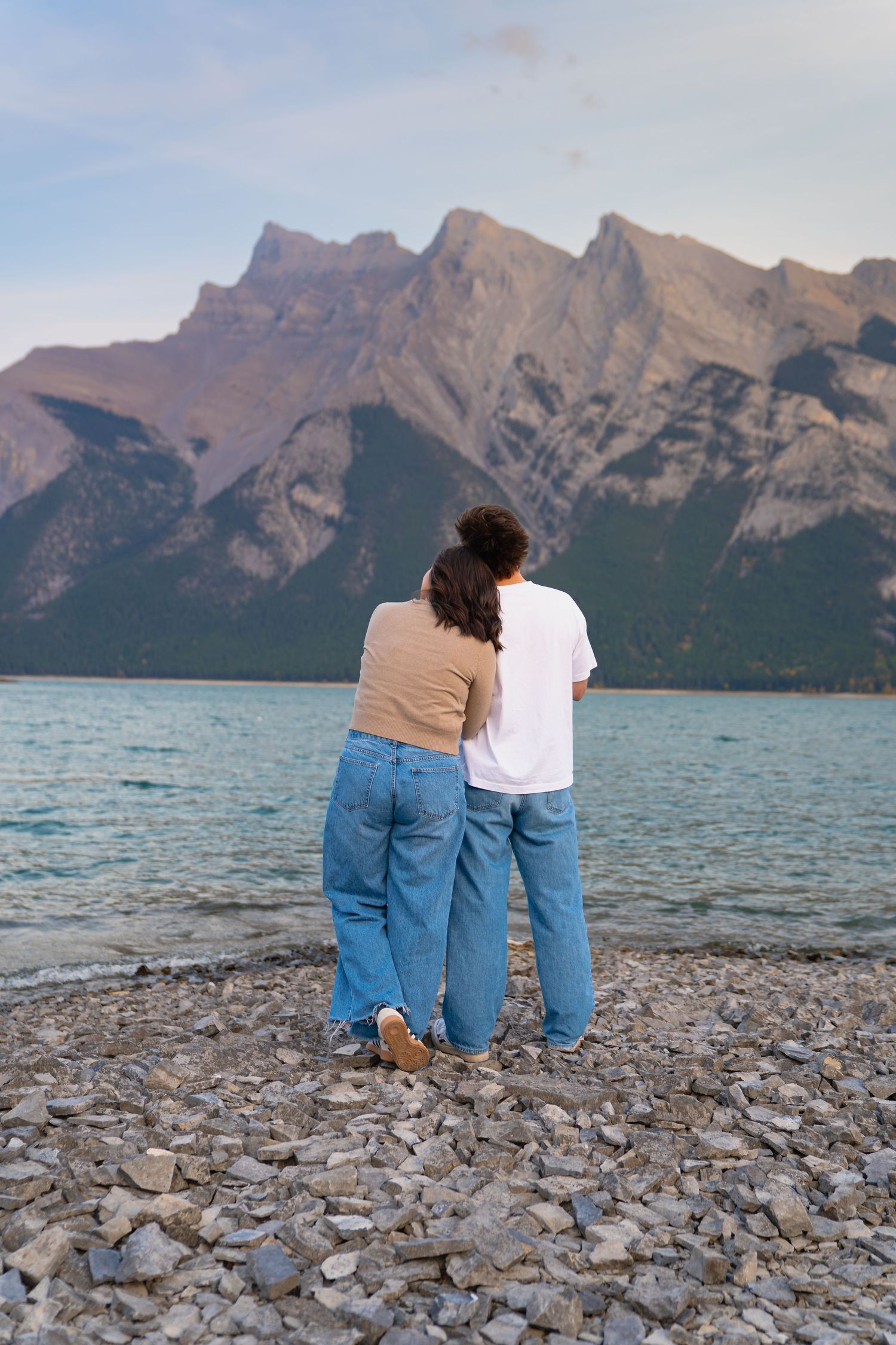 couple lean on each other and look off into the distance at lake Minnewanka in banff. a beautiful sunset with blue calm waters.