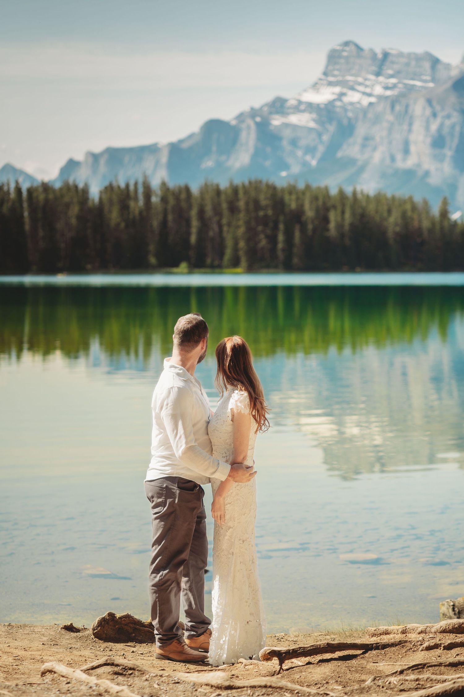 Bride and groom stand together by a calm Banff lake, looking out at dramatic Rocky Mountain peaks and reflections during a peaceful wedding