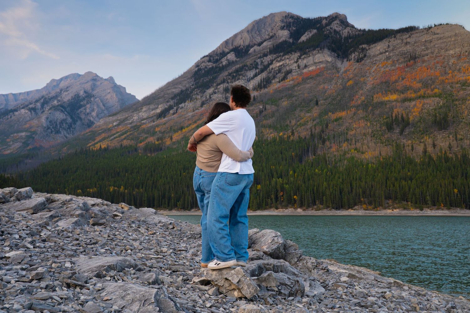 Couple embracing on rocky terrain at Lake Minnewanka in Banff, overlooking turquoise water and rugged mountains during a natural engagement