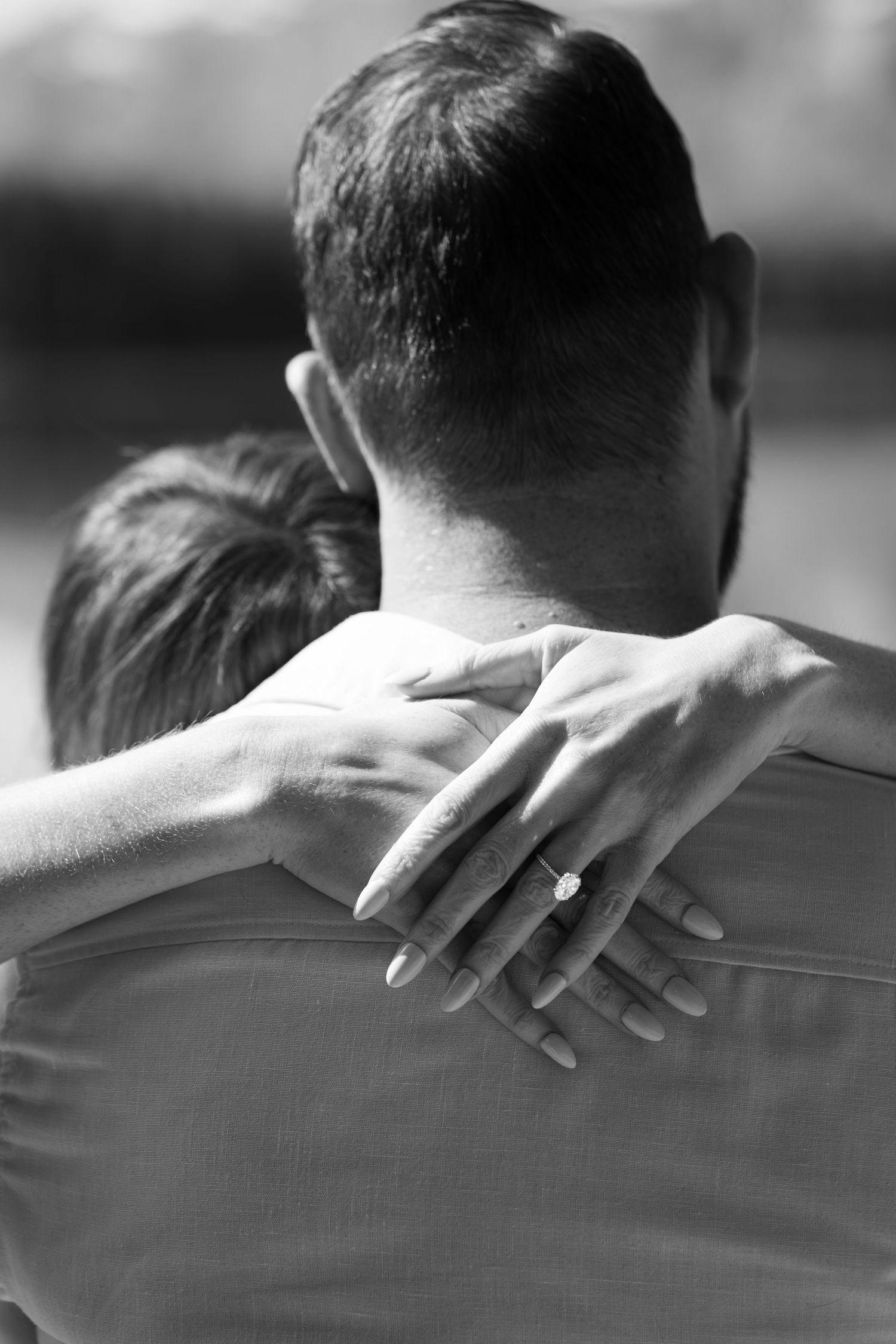 Hands wrapped around a partner’s shoulders,  a sparkling engagement ring in warm sunlight during a romantic outdoor moment. Black and white