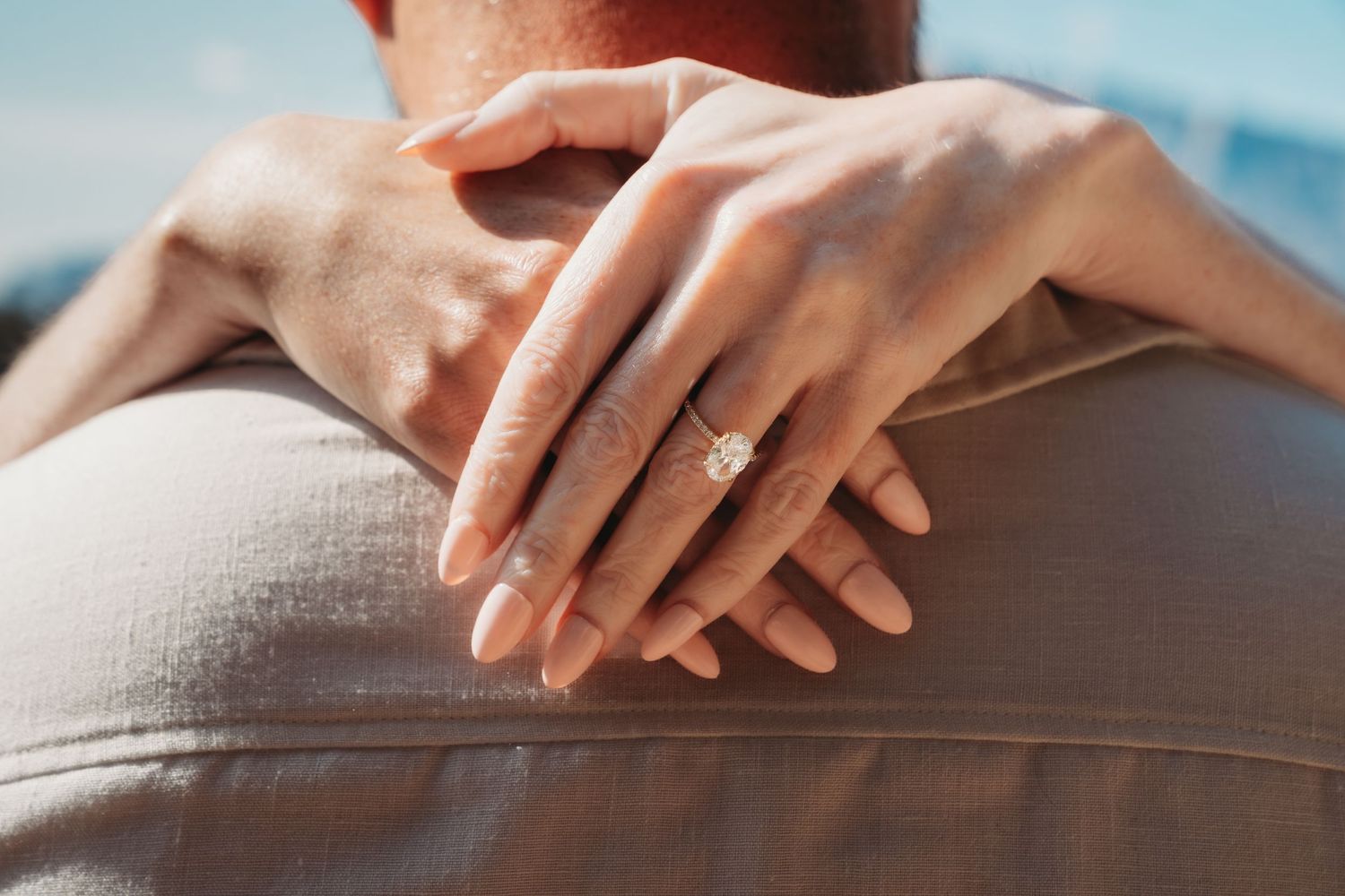 Hands wrapped around a partner’s shoulders, showcasing a sparkling engagement ring in warm sunlight during a romantic outdoor moment.