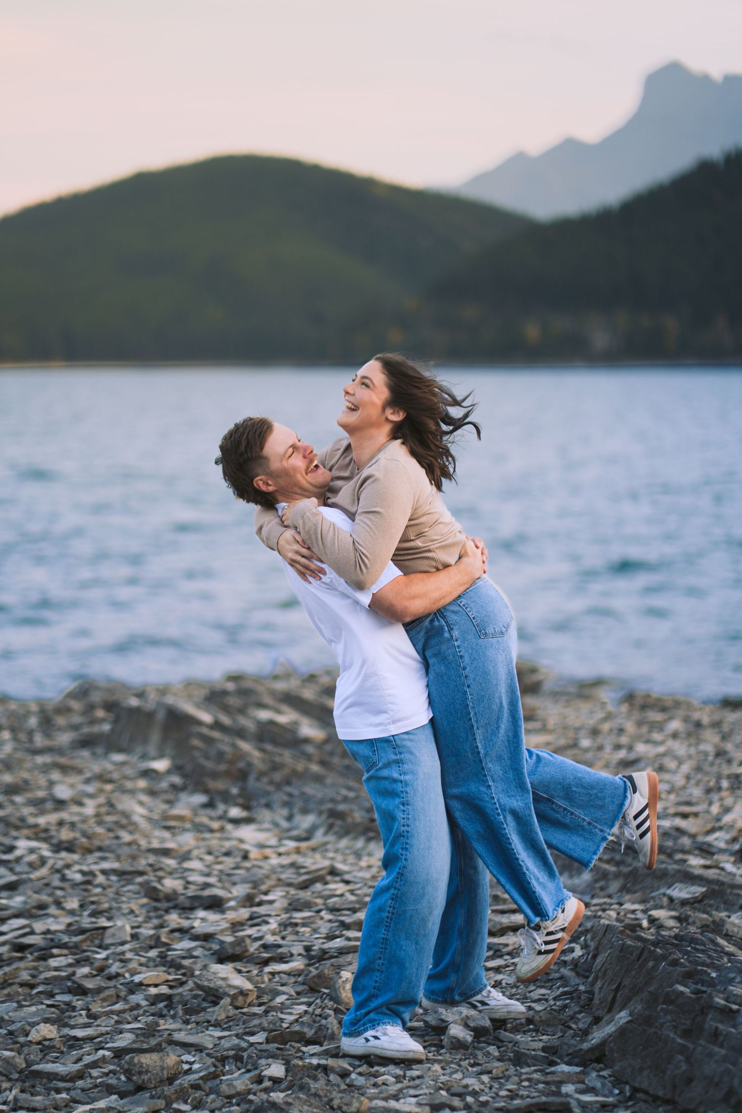 Fiancé lifts his partner while they laugh on the rocky shoreline at Lake Minnewanka in Banff, capturing a playful engagement moment.