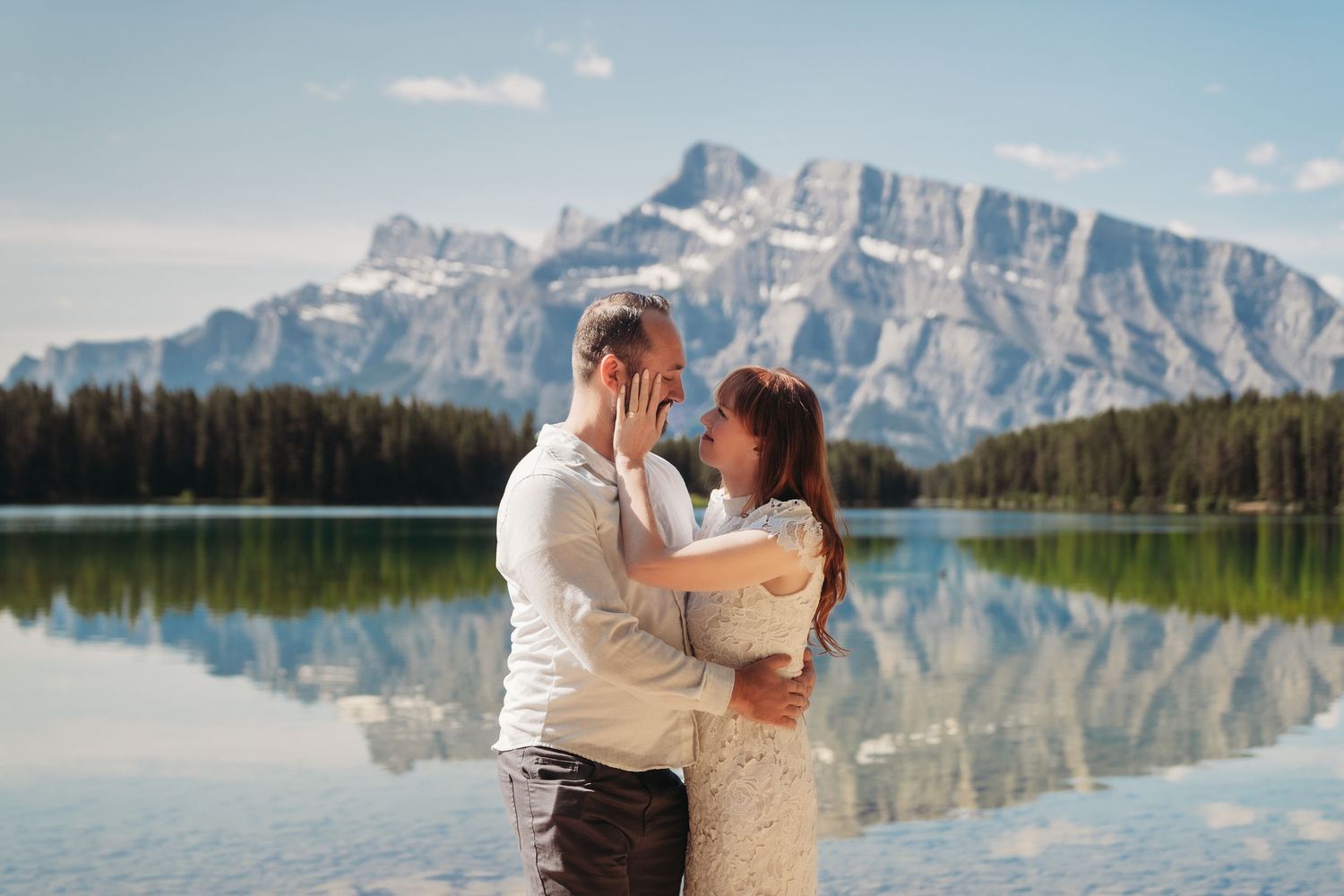 Bride and groom embrace by a clear mountain lake in Banff, surrounded by pine trees, reflections, and dramatic Rocky Mountain peaks.