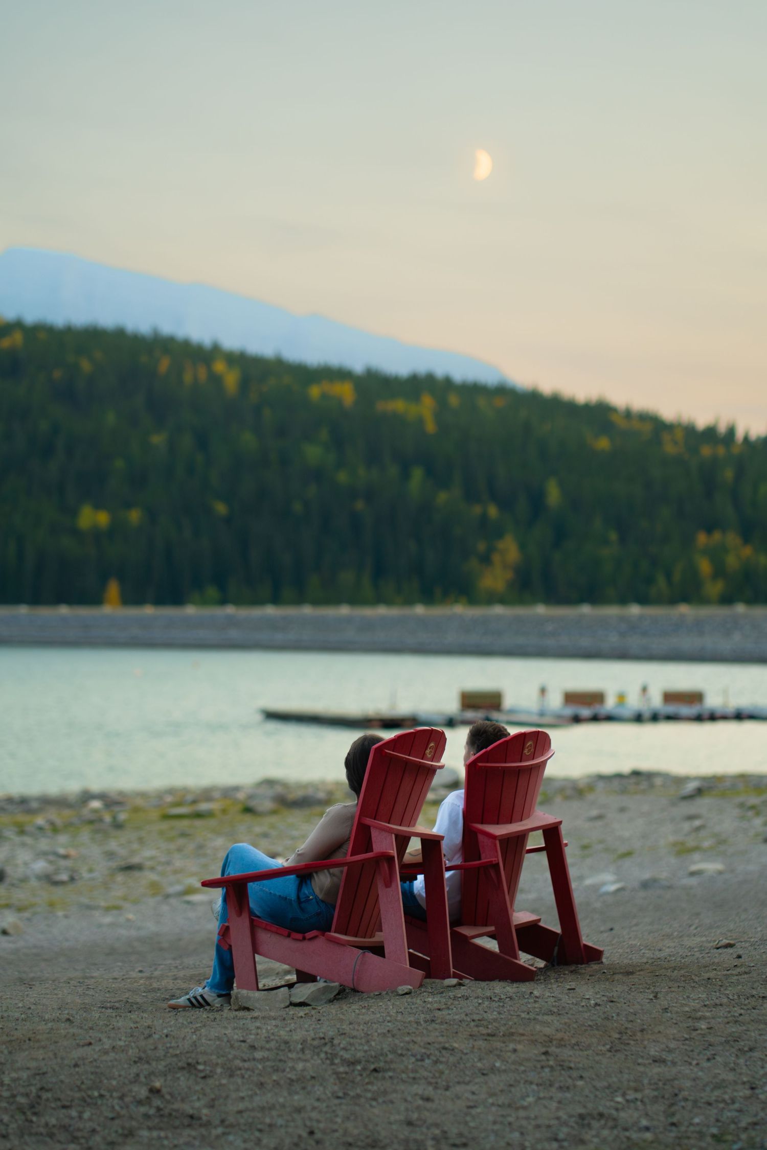 Couple sitting in red chairs beside Lake Minnewanka at dusk, looking toward the water and mountains during a peaceful engagement session.