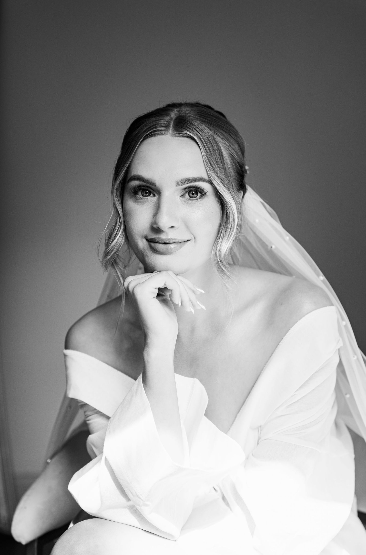 Bride poses in black and white with soft curls and veil, wearing a robe and looking confidently into the camera during wedding morning prep.