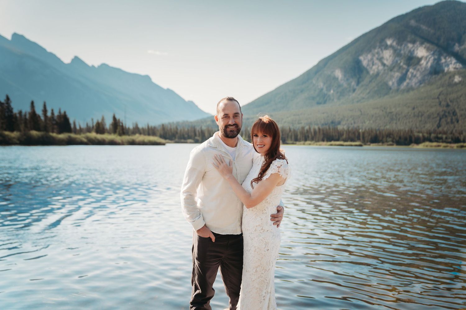 Bride and groom stand smiling by a calm mountain lake in Banff, surrounded by alpine peaks, evergreen trees, and clear blue summer skies.