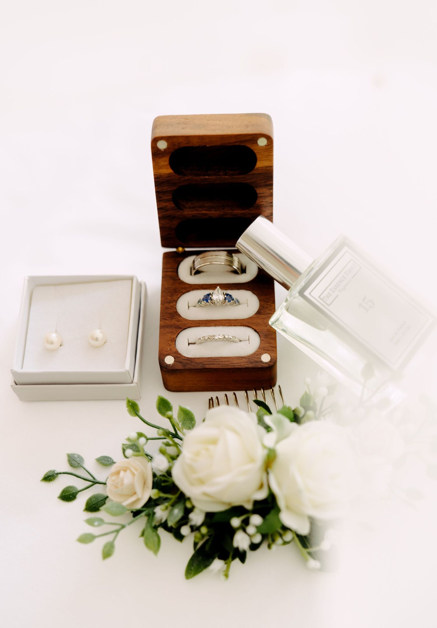 Wedding flatlay featuring rings in a wooden box, pearl earrings, perfume and white florals arranged on a clean white background.