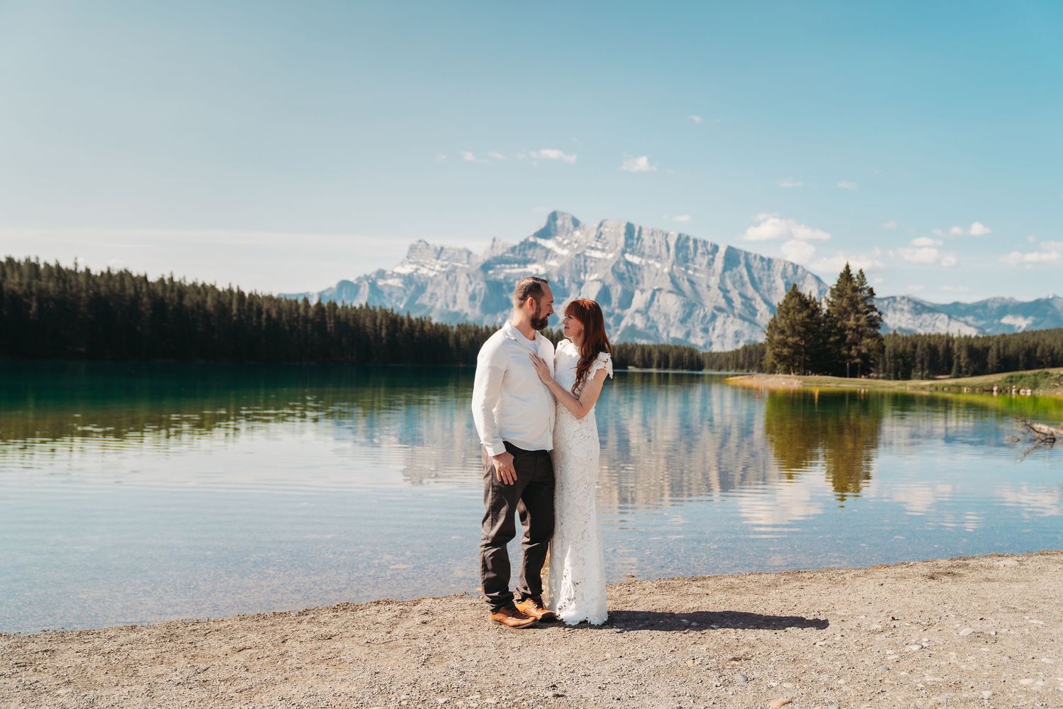Bride and groom stand by a calm mountain lake in Banff, sharing an intimate moment with clear reflections and rugged peaks in the background