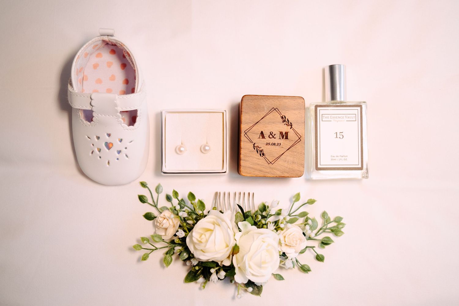 Wedding detail flatlay with baby shoe, pearl earrings, engraved ring box, perfume and floral hairpiece on white background.