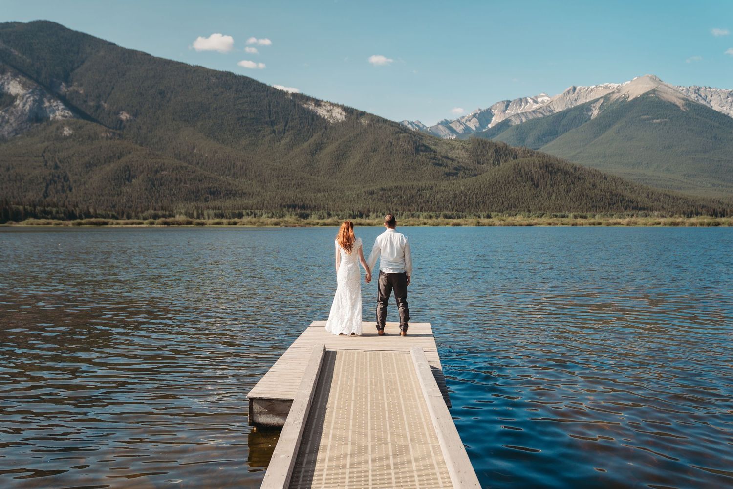 Couple stand on the end of a dock looking out at mount Rundle in Banff. overlooking a calm lake, surrounded by forested peaks.