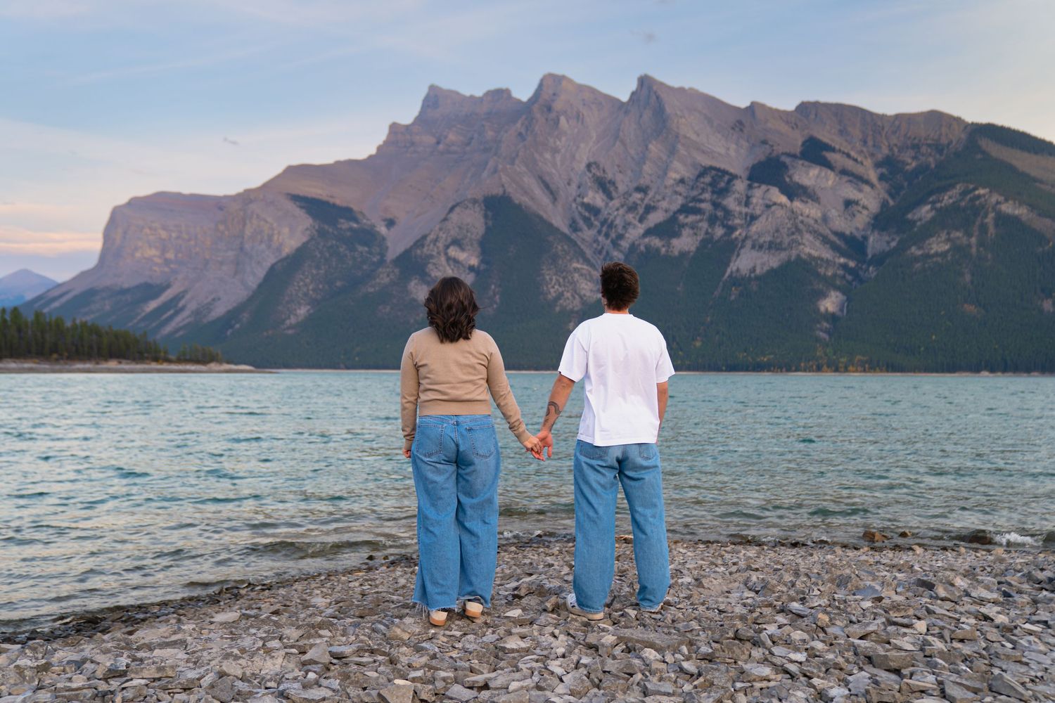 couple hold hands and look off into the distance at lake Minnewanka in banff turquoise water and rugged mountains during a loving moment