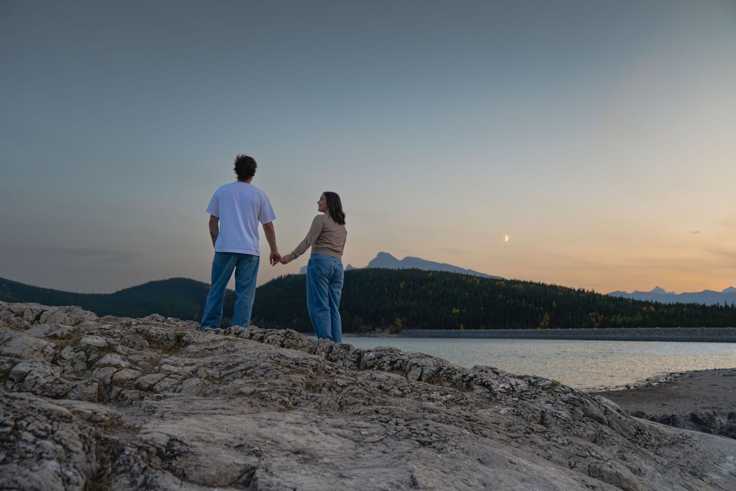 Couple holding hands while overlooking Lake Minnewanka at sunset in Banff,  rocky terrain with mountains and calm water in the background.