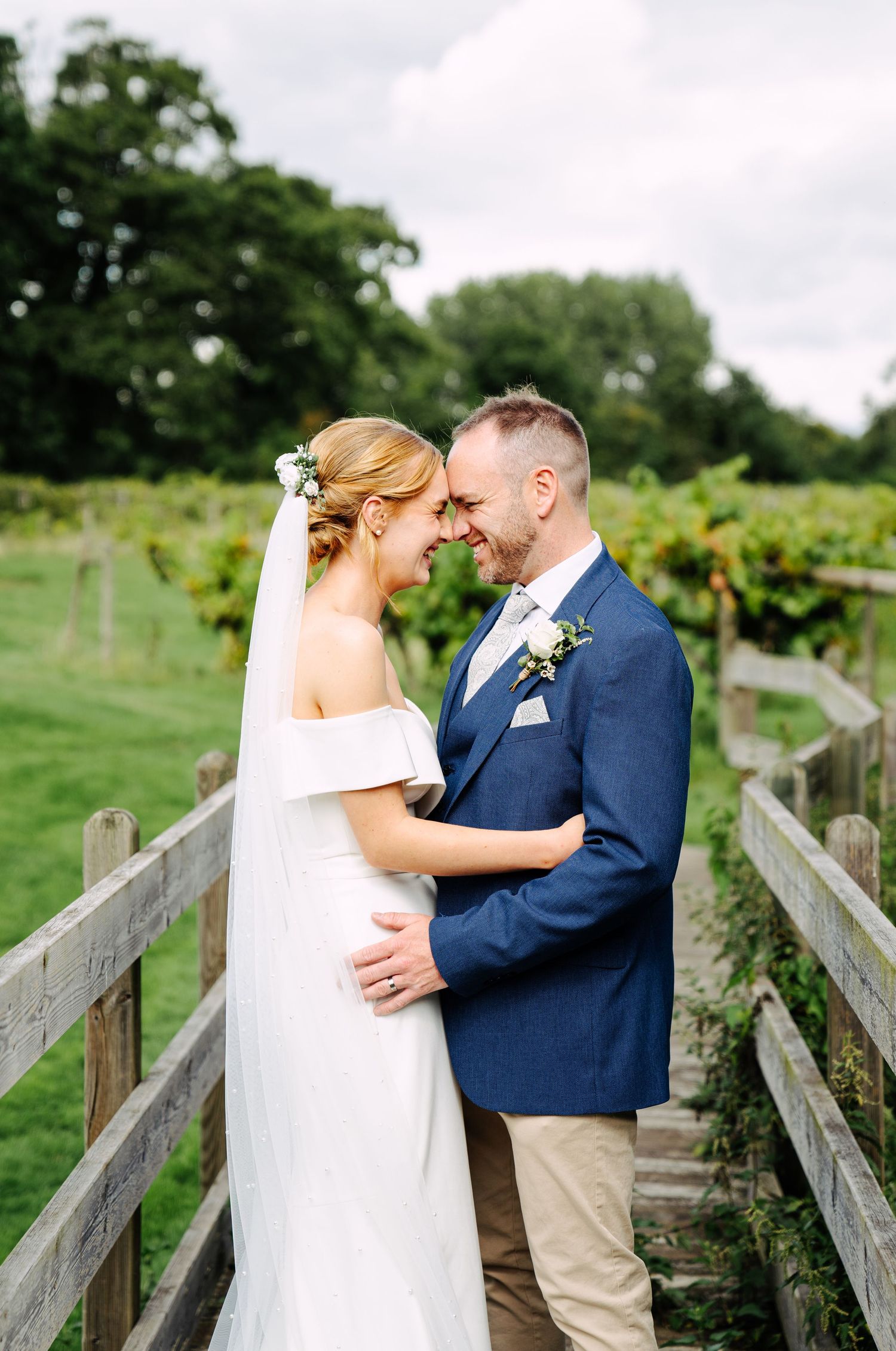 A posed photo of a bride and groom together on a wooden bridge. Their foreheads pressed together. Gazing into each other's eyes.