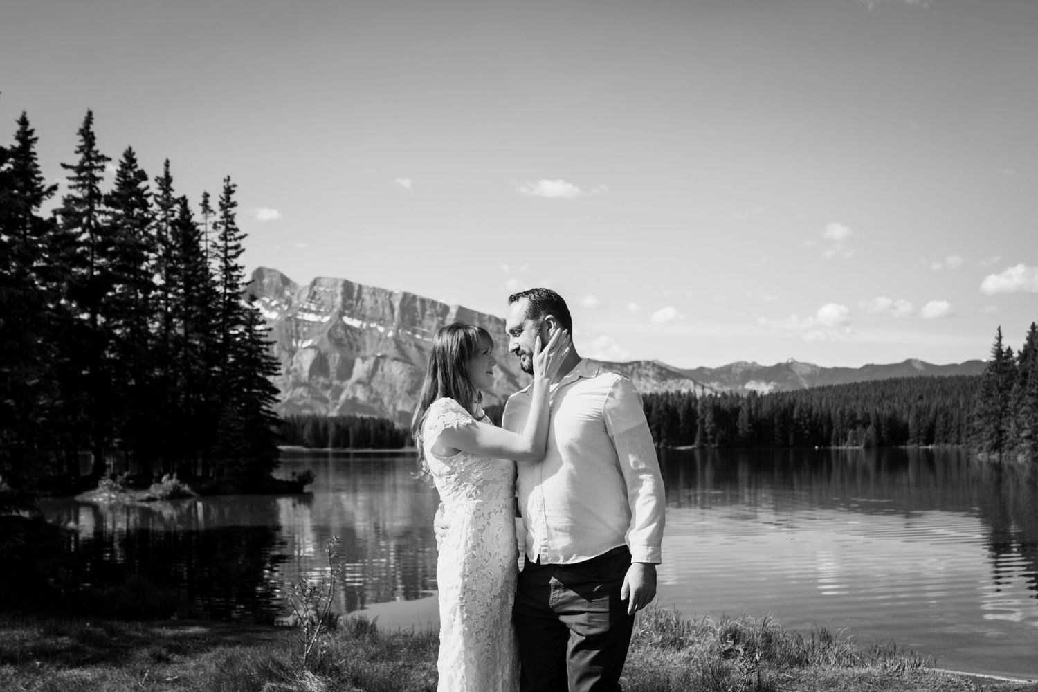 Bride and groom share a tender moment by a calm mountain lake in Banff, captured in black and white with dramatic peaks and evergreen trees