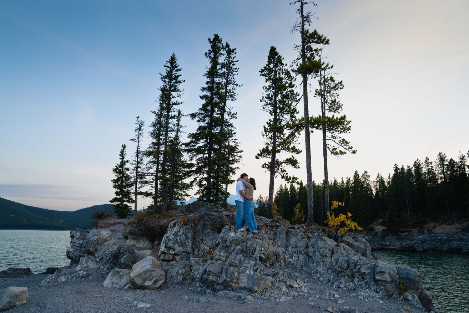 Couple embracing on a rocky shoreline at Lake Minnewanka in Banff, surrounded by tall pine trees and calm evening light during an engagement
