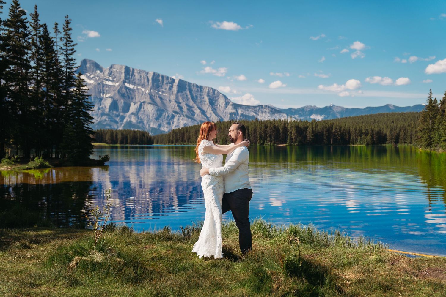Bride and groom embrace by a clear mountain lake in Banff, surrounded by pine trees, reflections, and dramatic Rocky Mountain peaks.