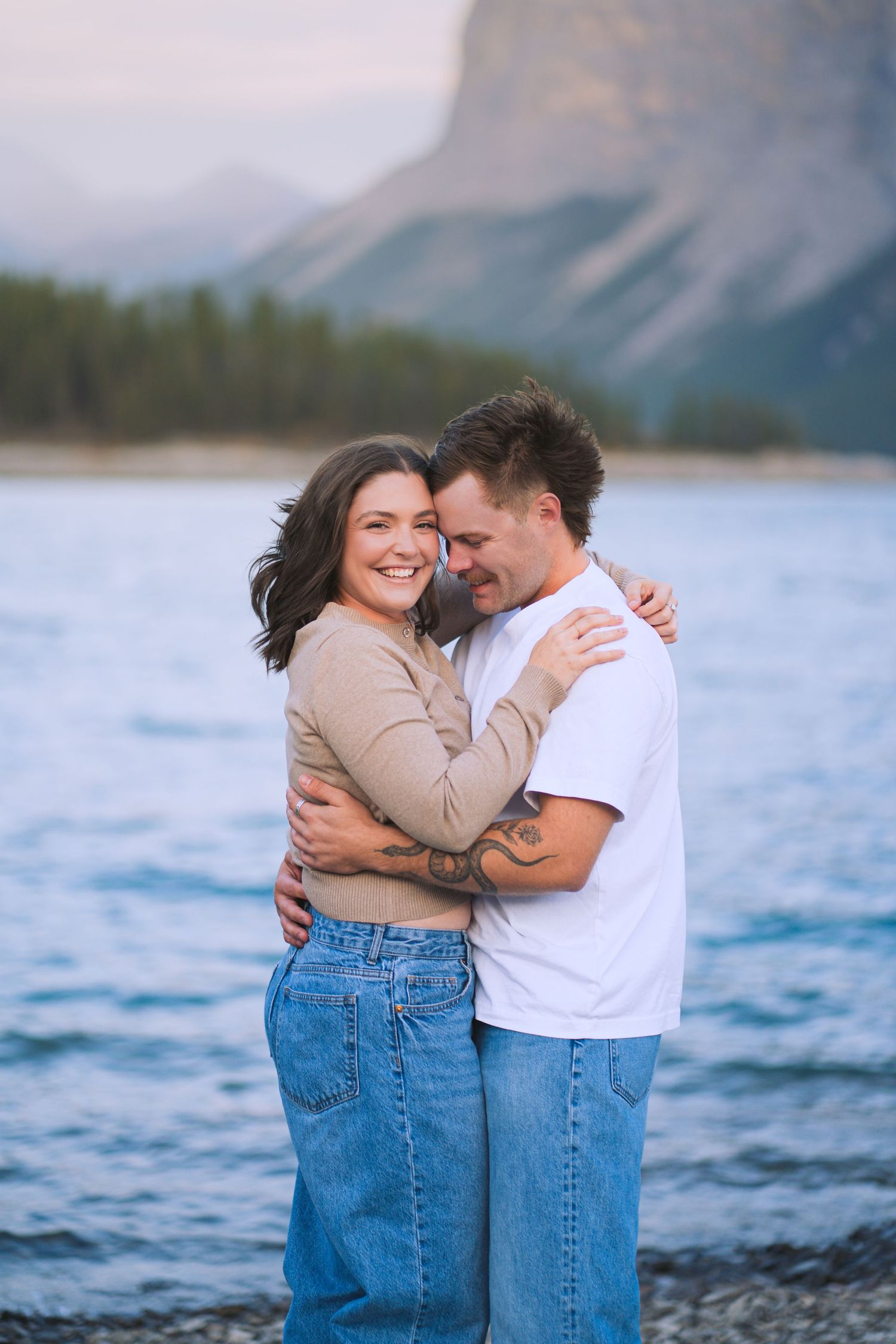 Couple embracing and smiling at Lake Minnewanka in Banff, with mountains and water behind them during a relaxed engagement shoot.