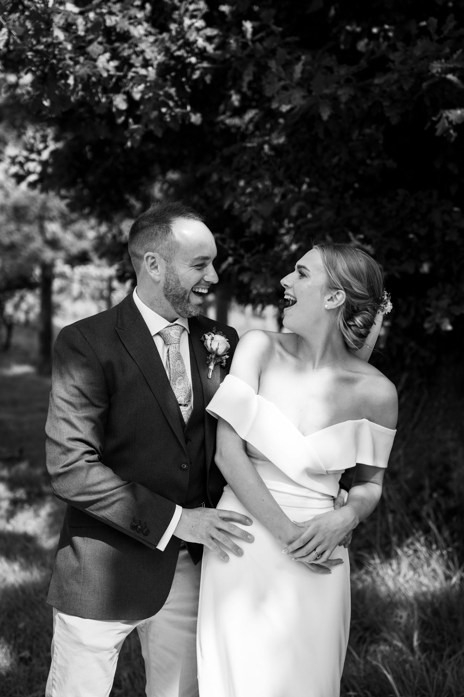 A candid portrait of a bride and groom sharing a happy moment together beneath leafy trees, captured in soft natural light. Black and white.